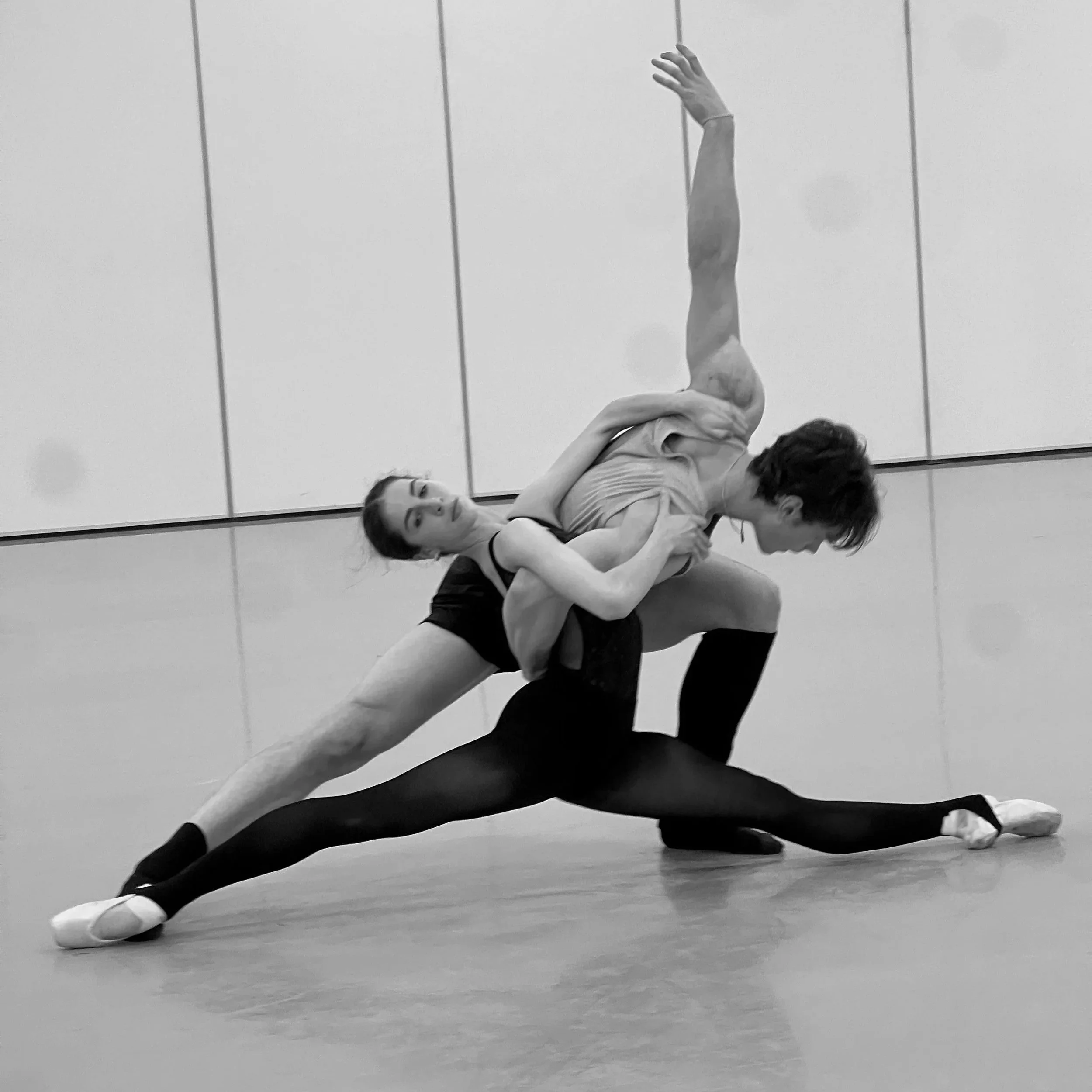 Two ballet dancers practicing together, one supporting the other in a stretch pose, on a dance studio floor.