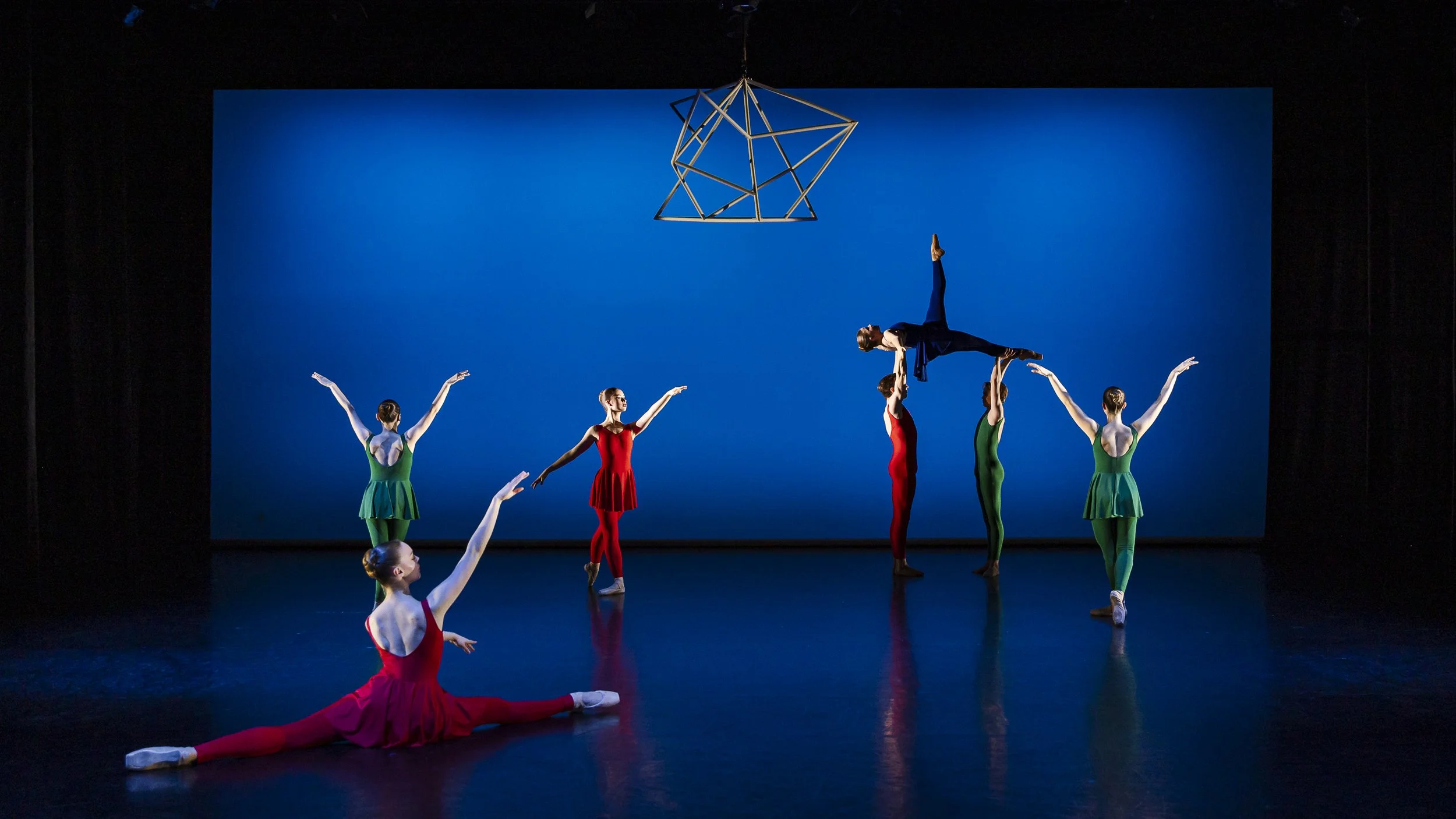 Ballet dancers practicing on stage, with one dancer being lifted horizontally by two others, and three dancers in green and red costumes in various poses, against a blue backdrop.