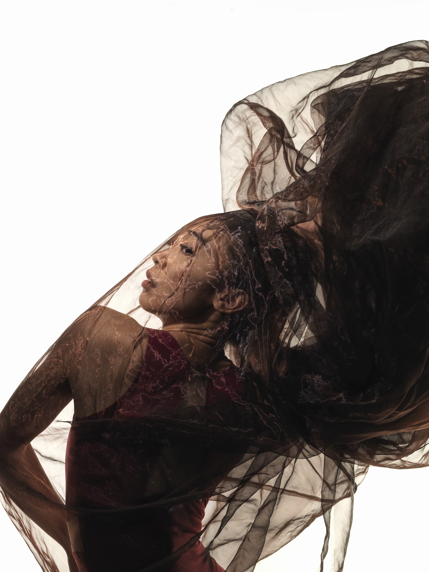 A woman is posing with sheer, flowing fabric draped over her head and shoulder, creating a dynamic, artistic composition.