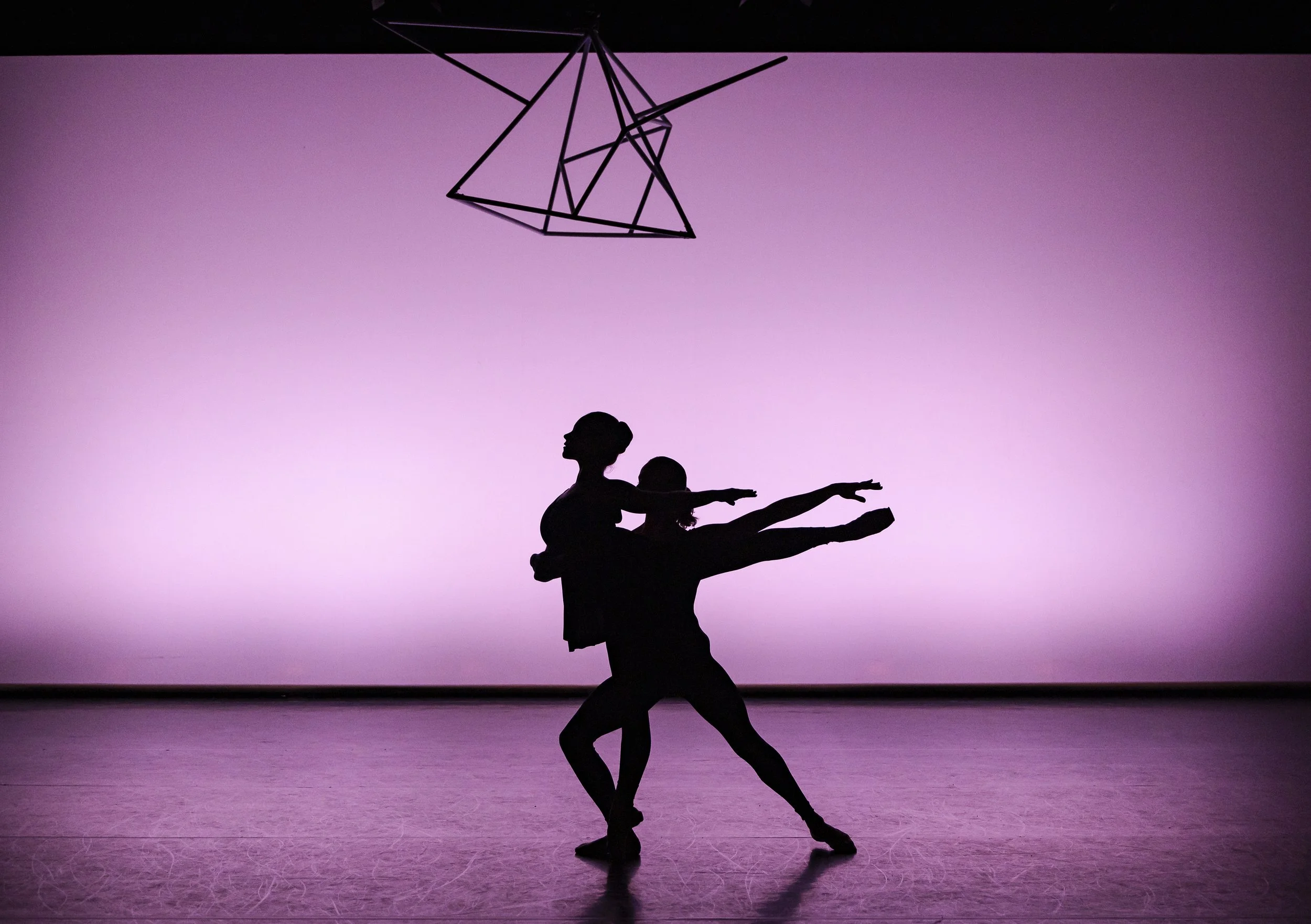 Silhouettes of two ballet dancers performing on stage with a pinkish background, and a geometric floral light fixture hanging above.