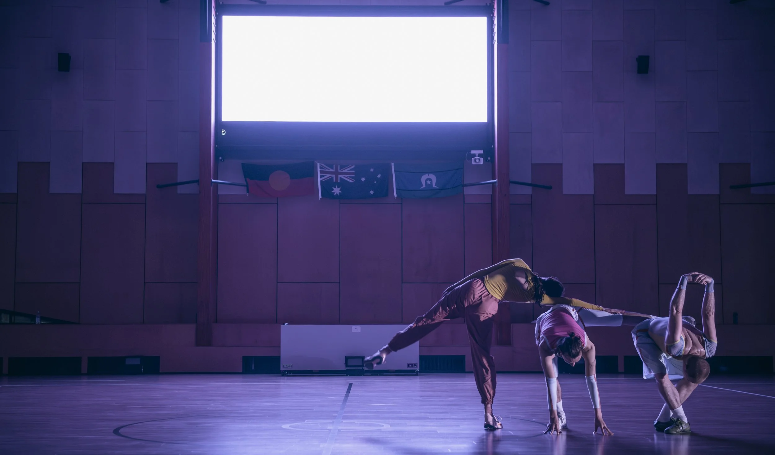 Three dancers in a gymnasium performing a choreographed move, with Australian, Aboriginal, and other flags hanging on the wall behind them.