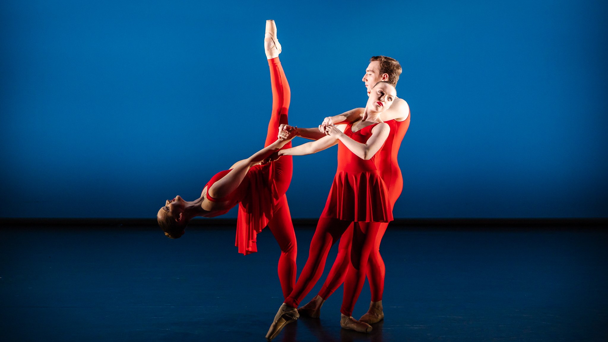 Two ballet dancers in red costumes performing on stage against a dark blue background.