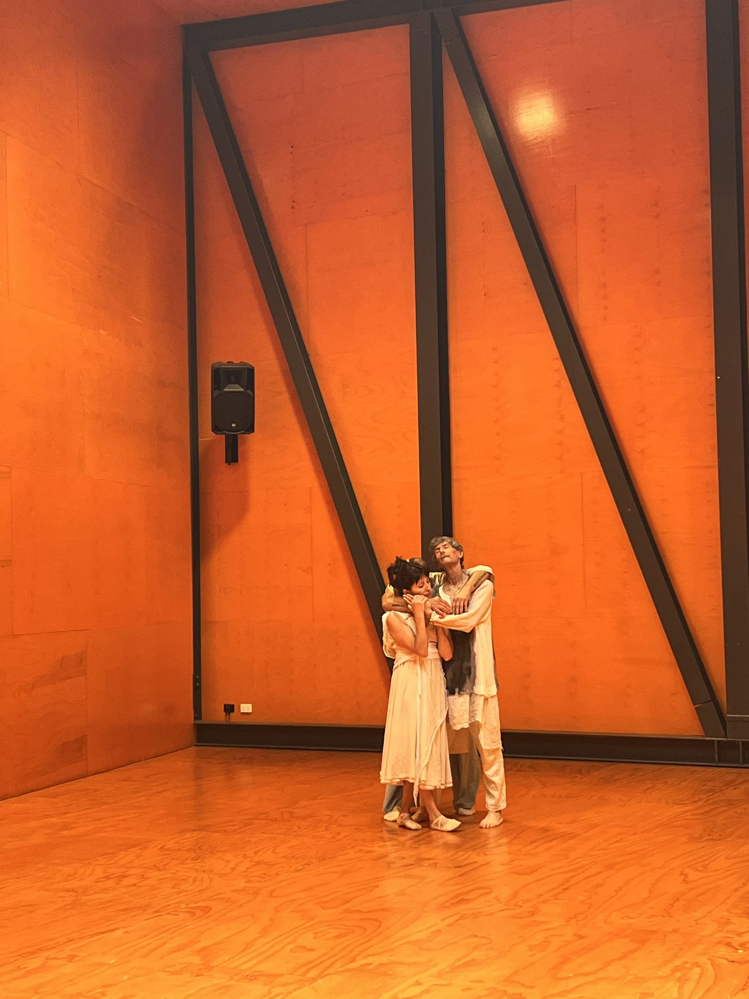 Two performers on stage hug each other against a large orange wall with black structural accents in a theater.