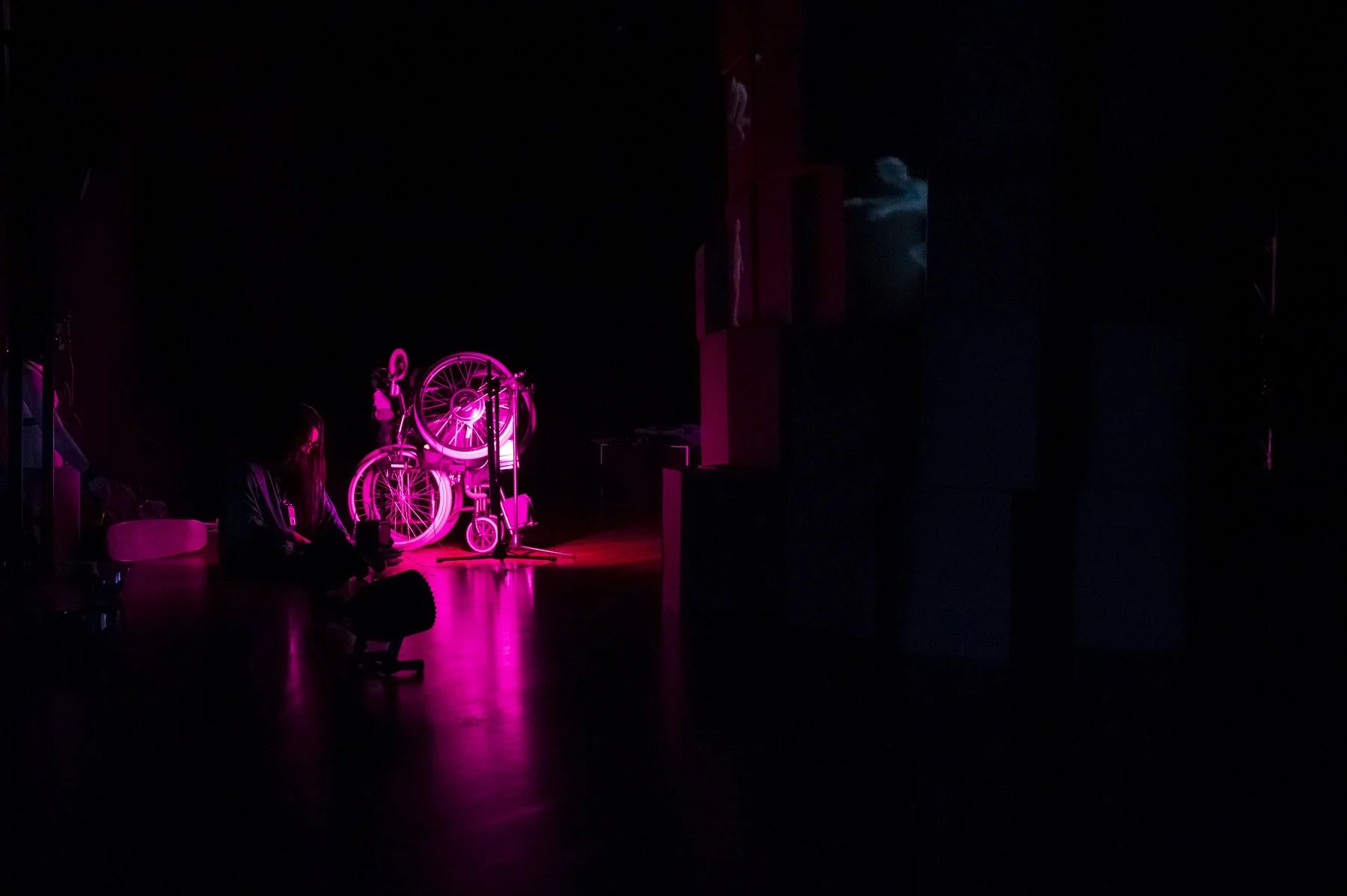 Dark room illuminated by pink light, with a woman sitting on the floor near pink bicycle sculptures and a microphone stand.