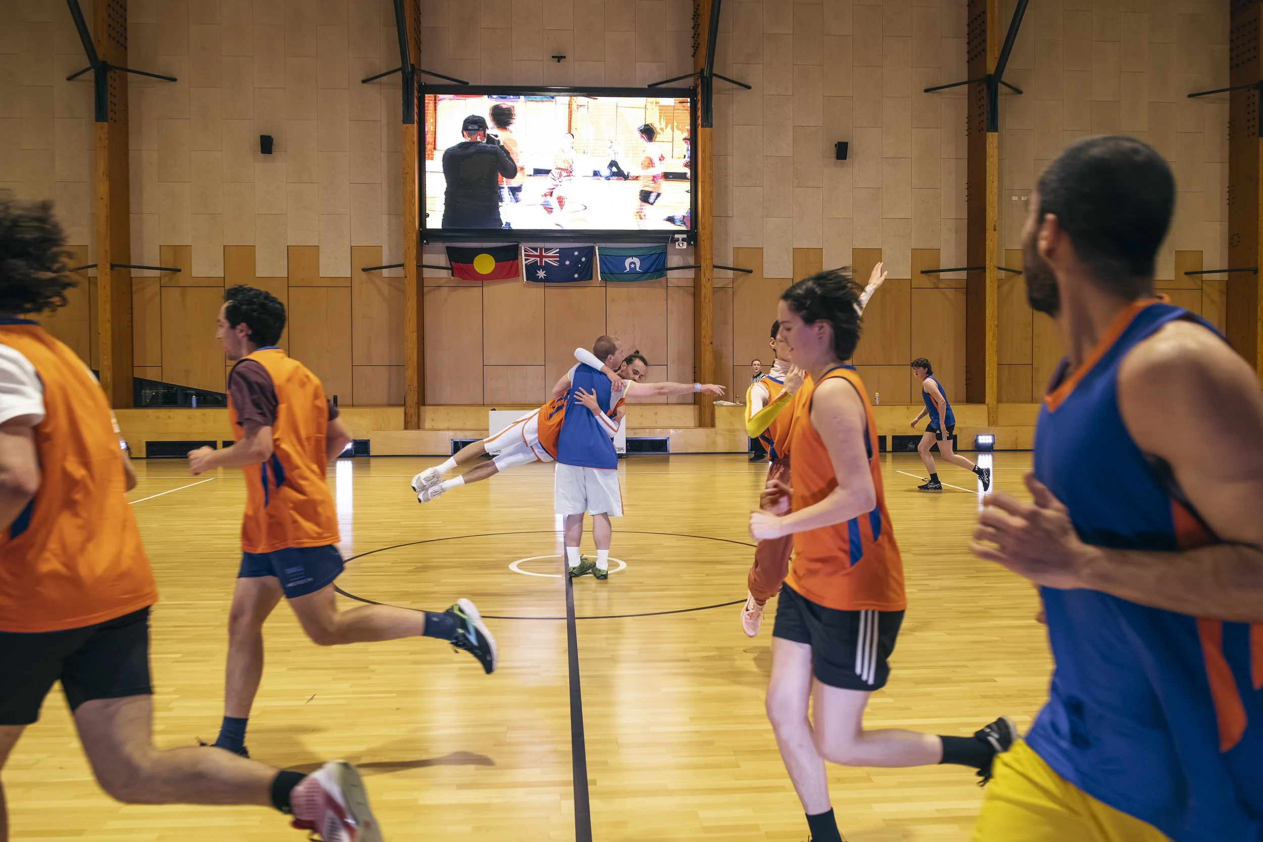 People playing basketball indoors, with two players in blue uniforms and others in orange, some running and jumping, in a gymnasium with flags and a large screen in the background.