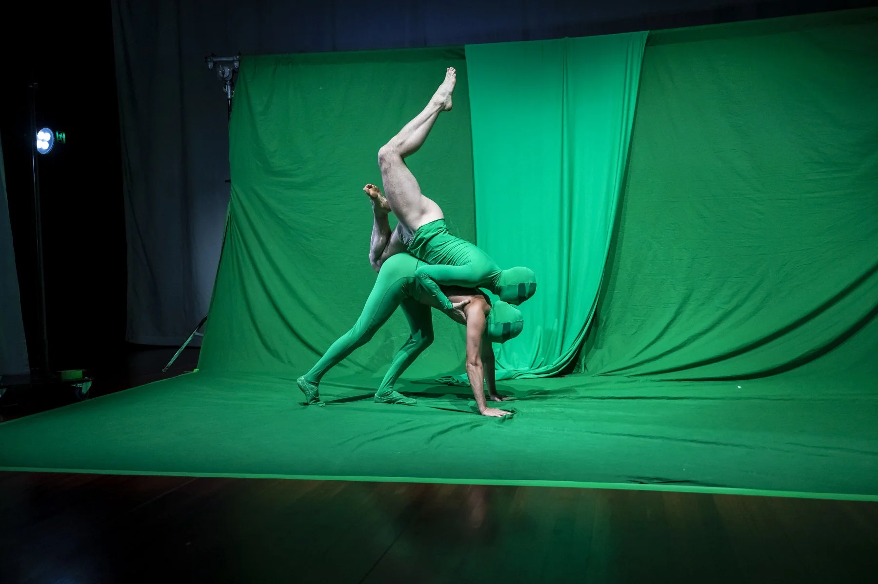 Two performers in green costumes and head coverings perform an acrobatic pose on a green backdrop and floor.