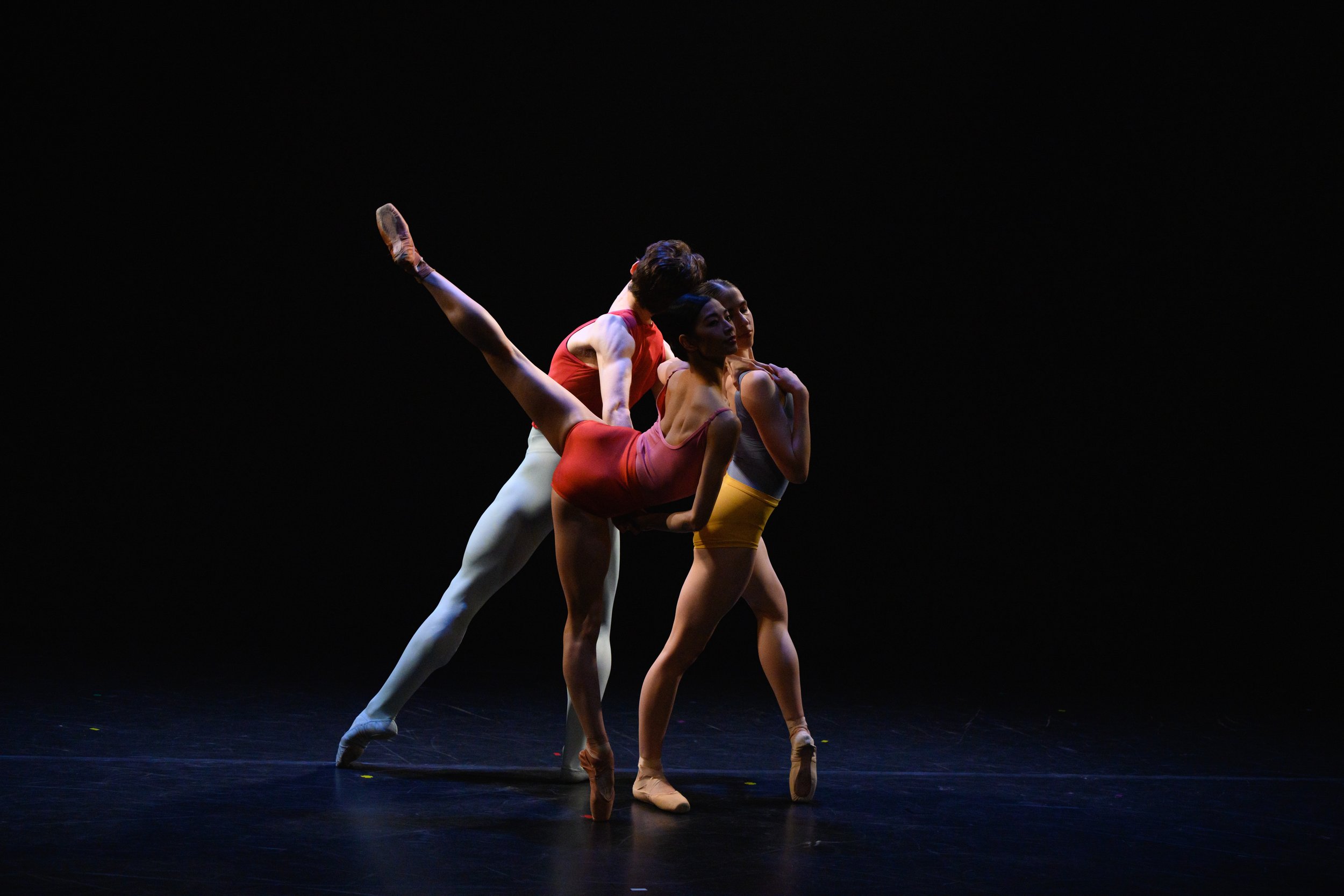 Three ballet dancers perform on stage with a dark background, wearing colorful costumes, one lifting another in an elegant pose.