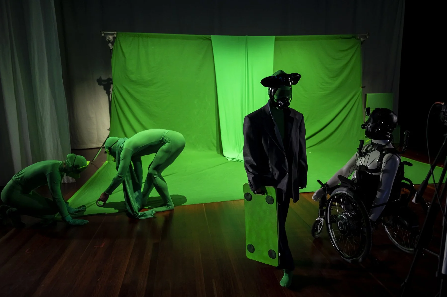 Studio scene with green screen backdrop, two human figures in bodysuits adjusting the setup, and a humanoid robot with a helmet and suit standing in front of the green screen.