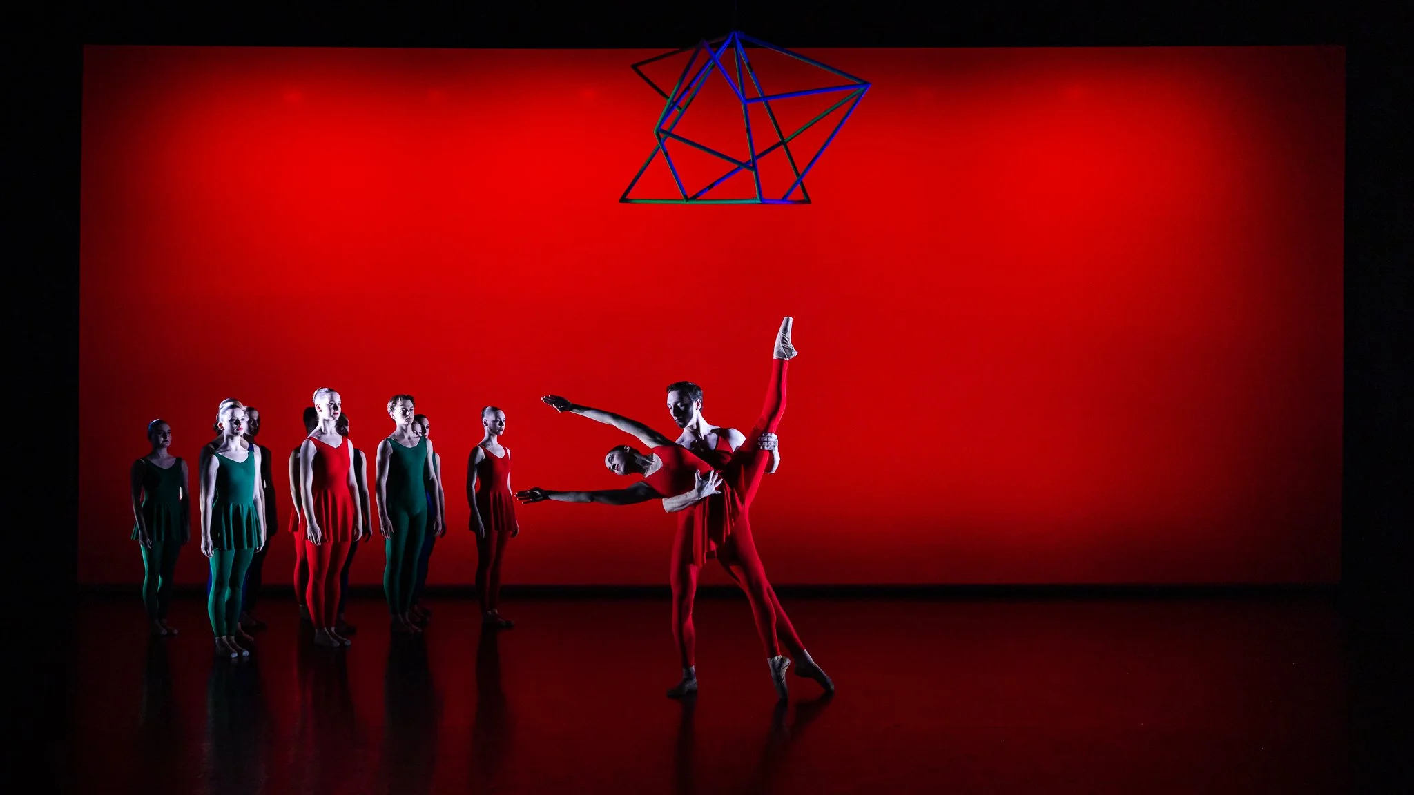 Ballet dancers performing on stage with a red background and geometric art hanging above, with some dancers in green and others in red costumes.