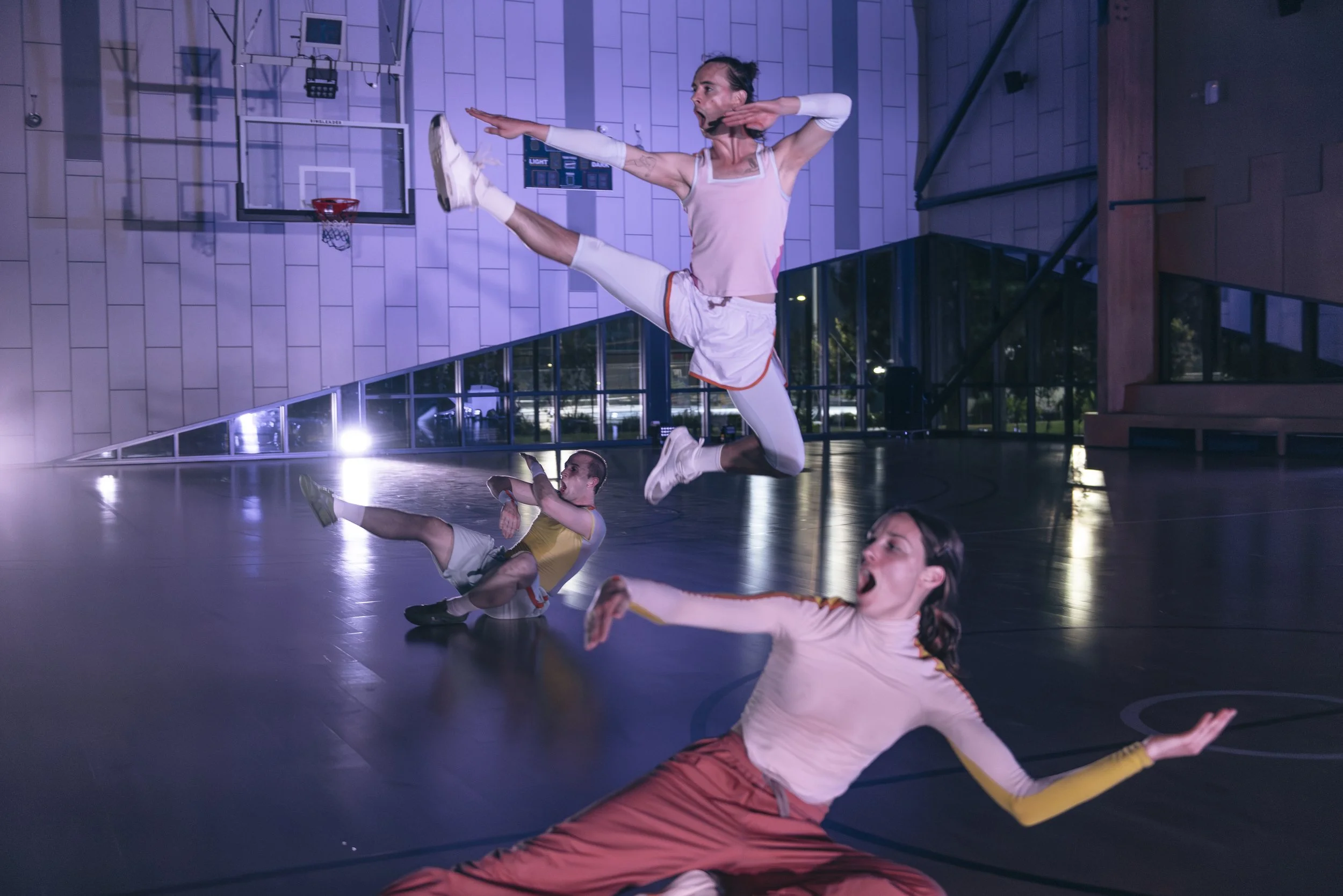 Three dancers in colorful outfits performing acrobatic dance moves on an indoor basketball court with a hoop and bright lights.