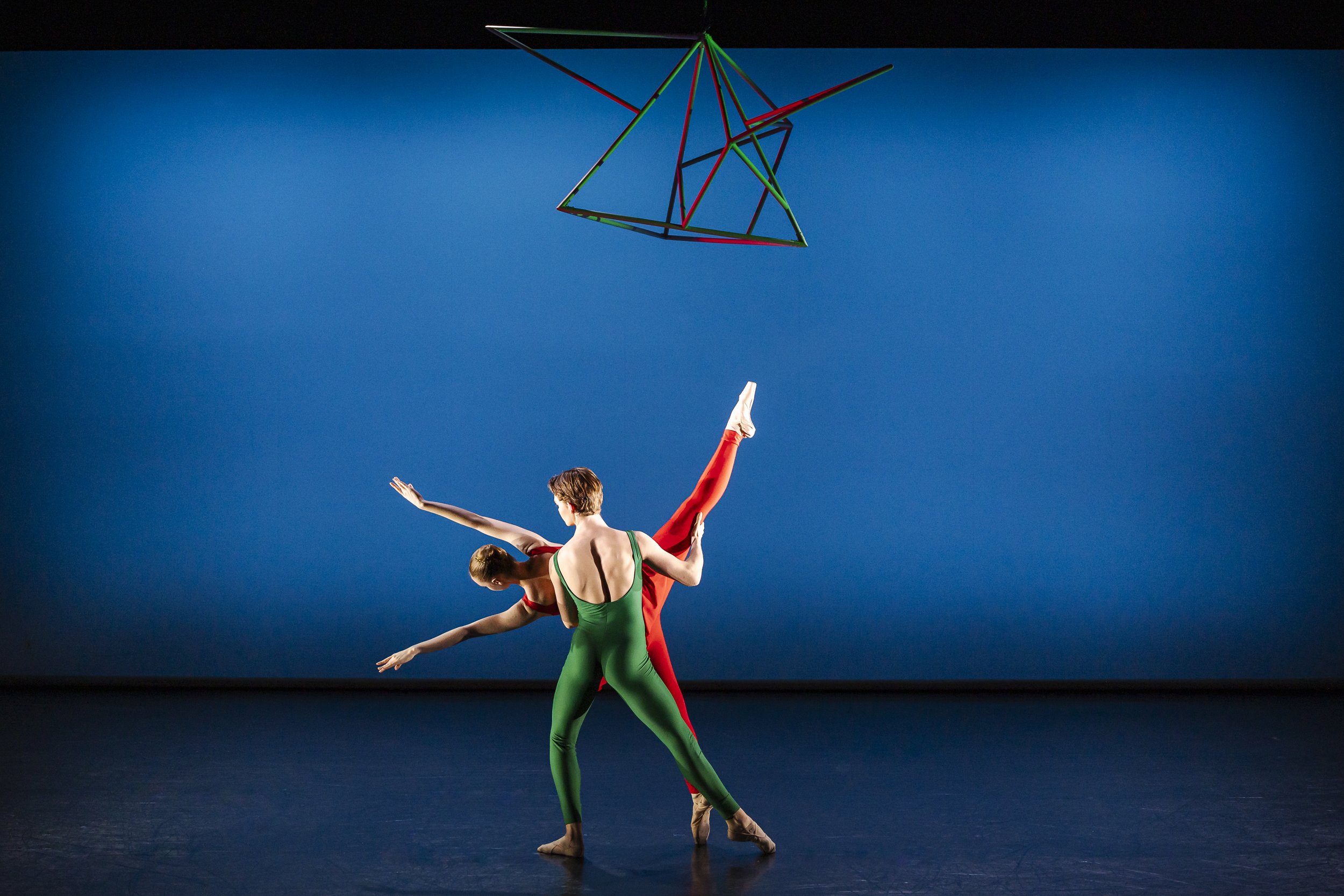 Two ballet dancers, one in a green costume and the other in a red costume, performing a dance move on stage with a blue background and a geometric hanging structure above them.