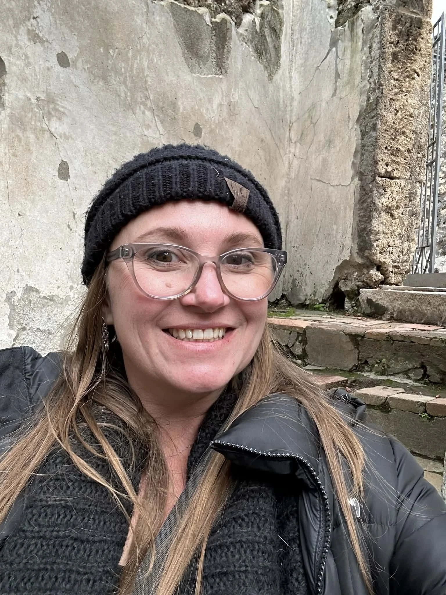 A woman with long blonde hair, glasses, and a black beanie smiling outdoors in front of an old stone wall and steps.