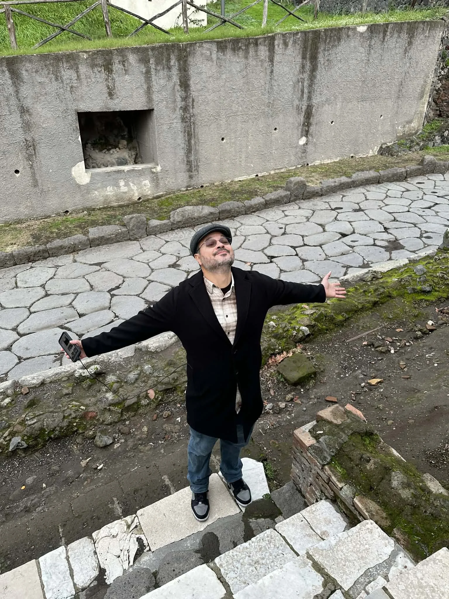A man with glasses and a beard, wearing a black jacket, jeans, and sneakers, stands with arms open at the bottom of a stone staircase outdoors. He is smiling and looking up, with a historic stone wall behind him and greenery above.