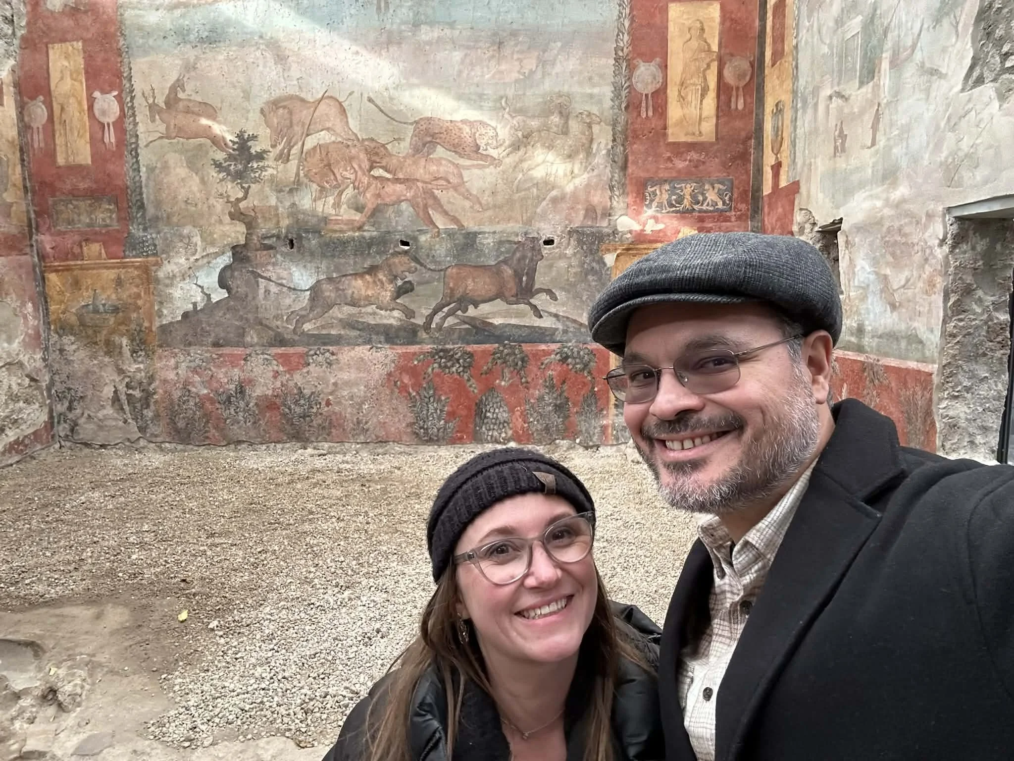 Two people taking a selfie in front of ancient Roman wall paintings depicting animals and figures, with a man wearing a gray cap and glasses and a woman with glasses and brown hair.