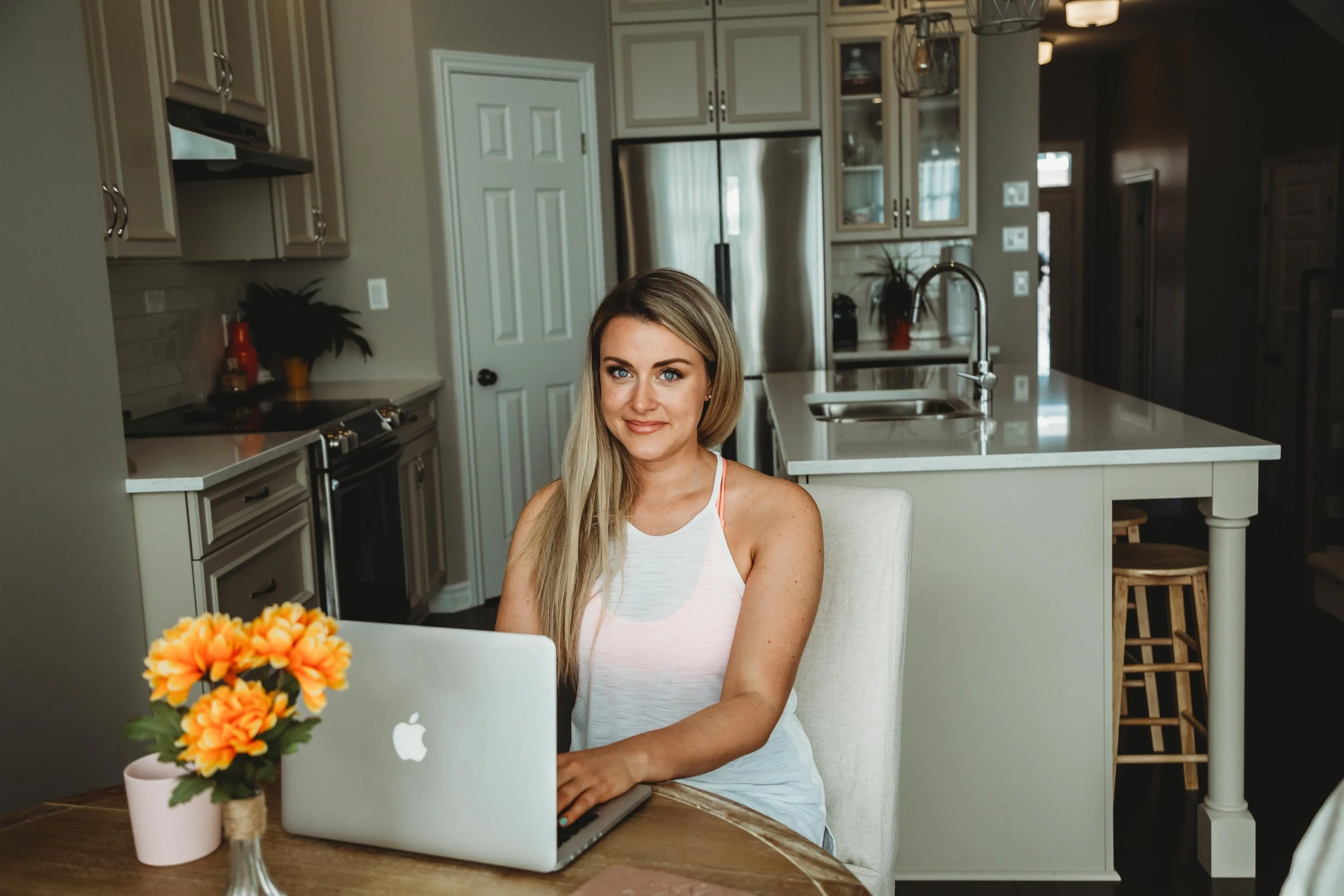 A woman with long blonde hair sitting at a dining table with a silver MacBook laptop and a pink flowerpot with orange flowers, in a modern kitchen with beige cabinets and stainless steel appliances.