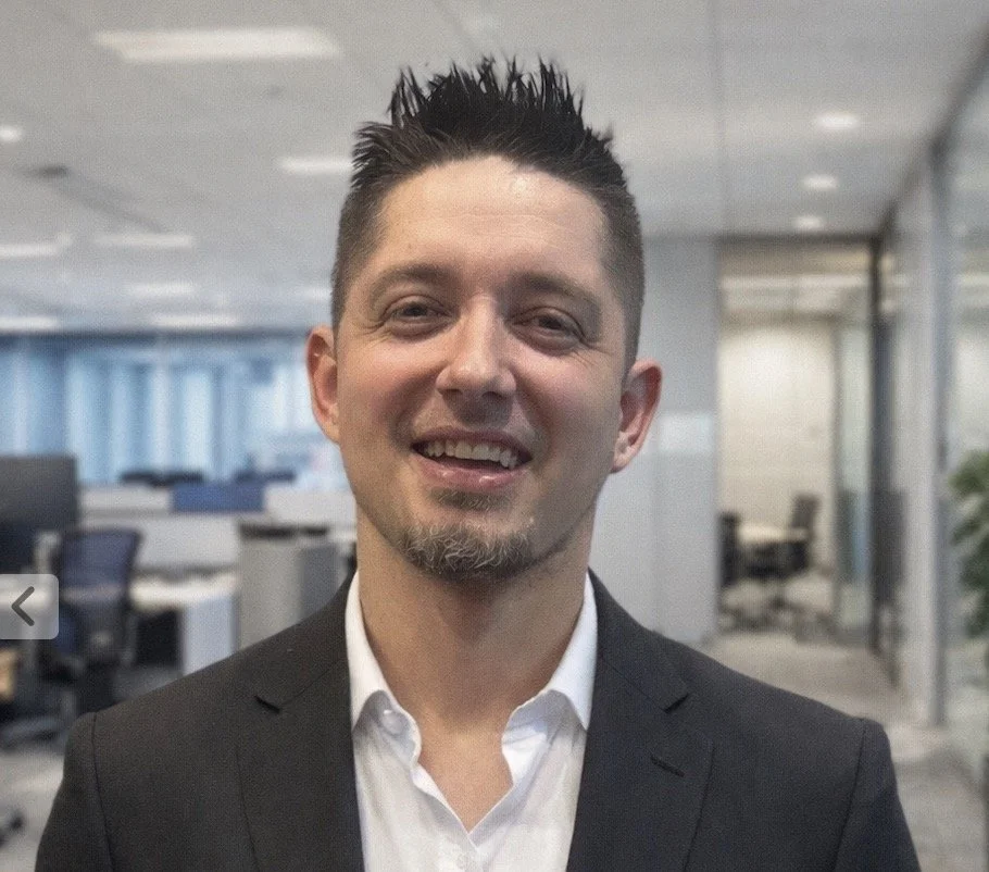 A man with spiked black hair, a goatee, and a mustache, dressed in a black suit and white shirt, smiling in a modern office setting.