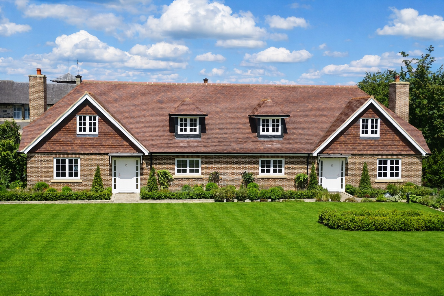 A large brick house with a red shingle roof, white doors, and multiple windows, surrounded by well-maintained grass and garden with small shrubs and trees under a blue sky with white clouds.