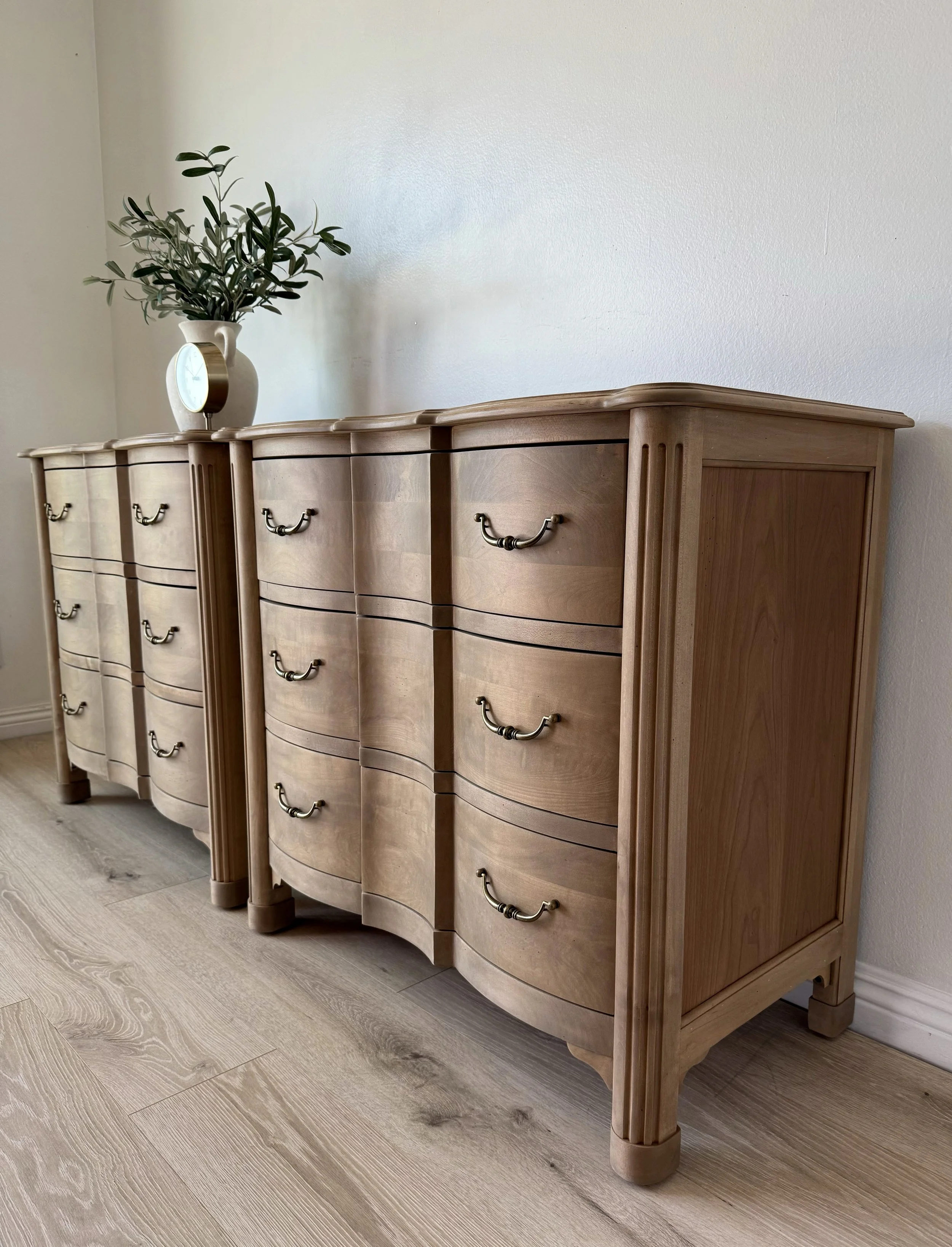 A wooden dresser with curved drawers and metal handles, topped with a white vase with green foliage and a small clock, placed against a plain white wall.