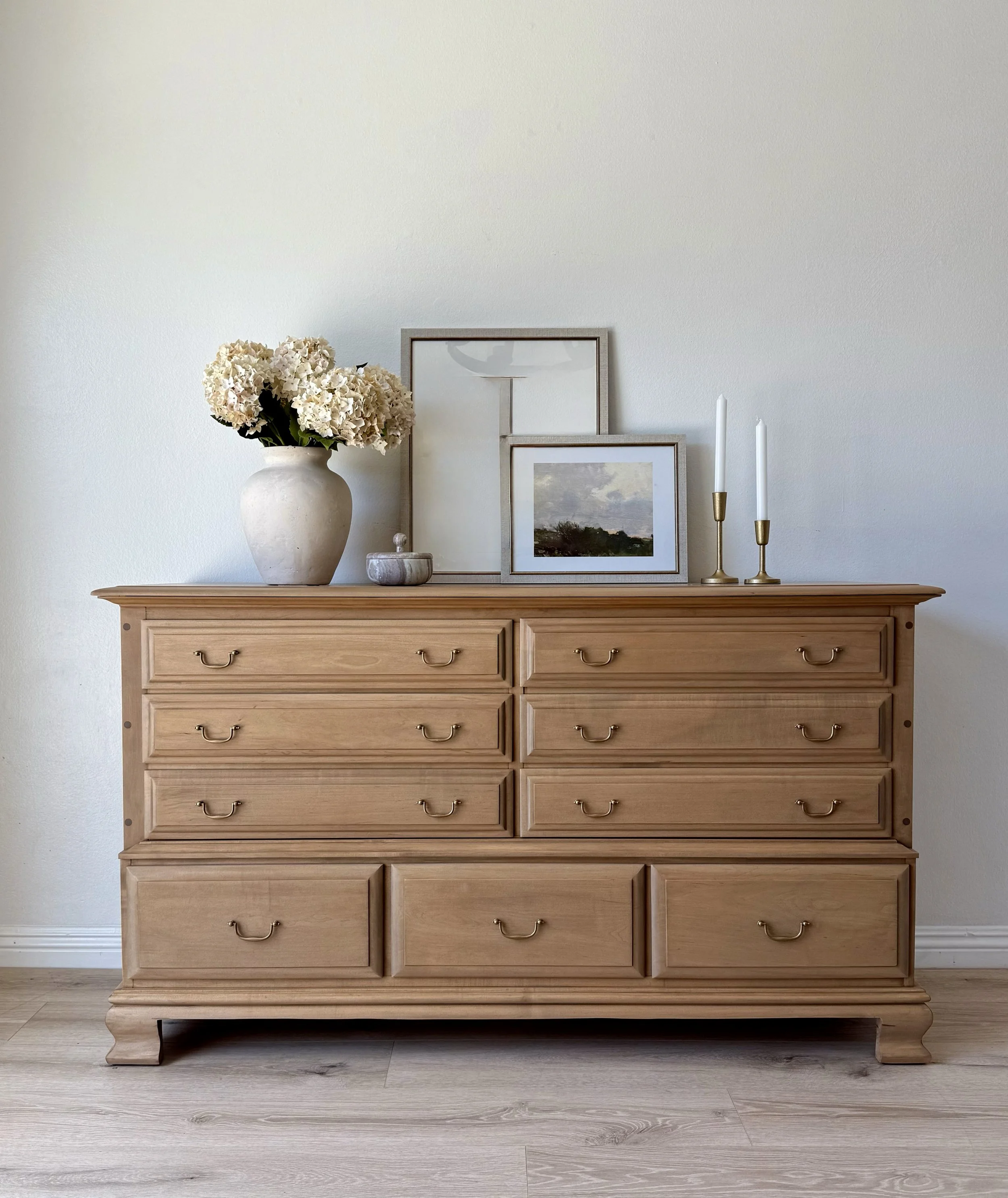A light wood dresser with eight small drawers and three larger drawers at the bottom, topped with a large white vase of hydrangeas, two picture frames, a small decorative object, and two white candlesticks with candles.