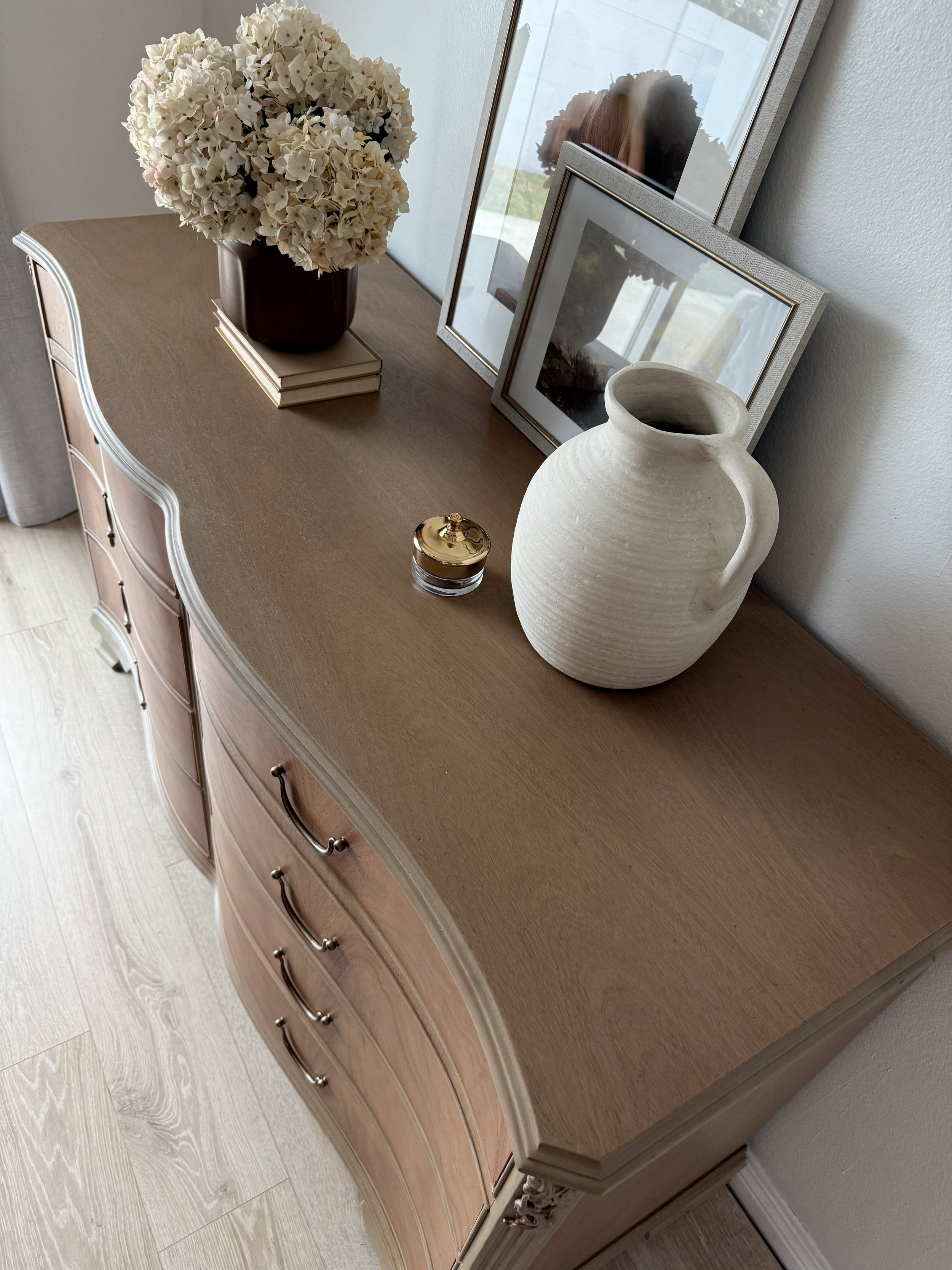 A wooden dresser decorated with a bouquet of white hydrangeas in a dark vase, two framed pictures, a white ceramic vase, and a small jar with a gold lid, placed near a window.