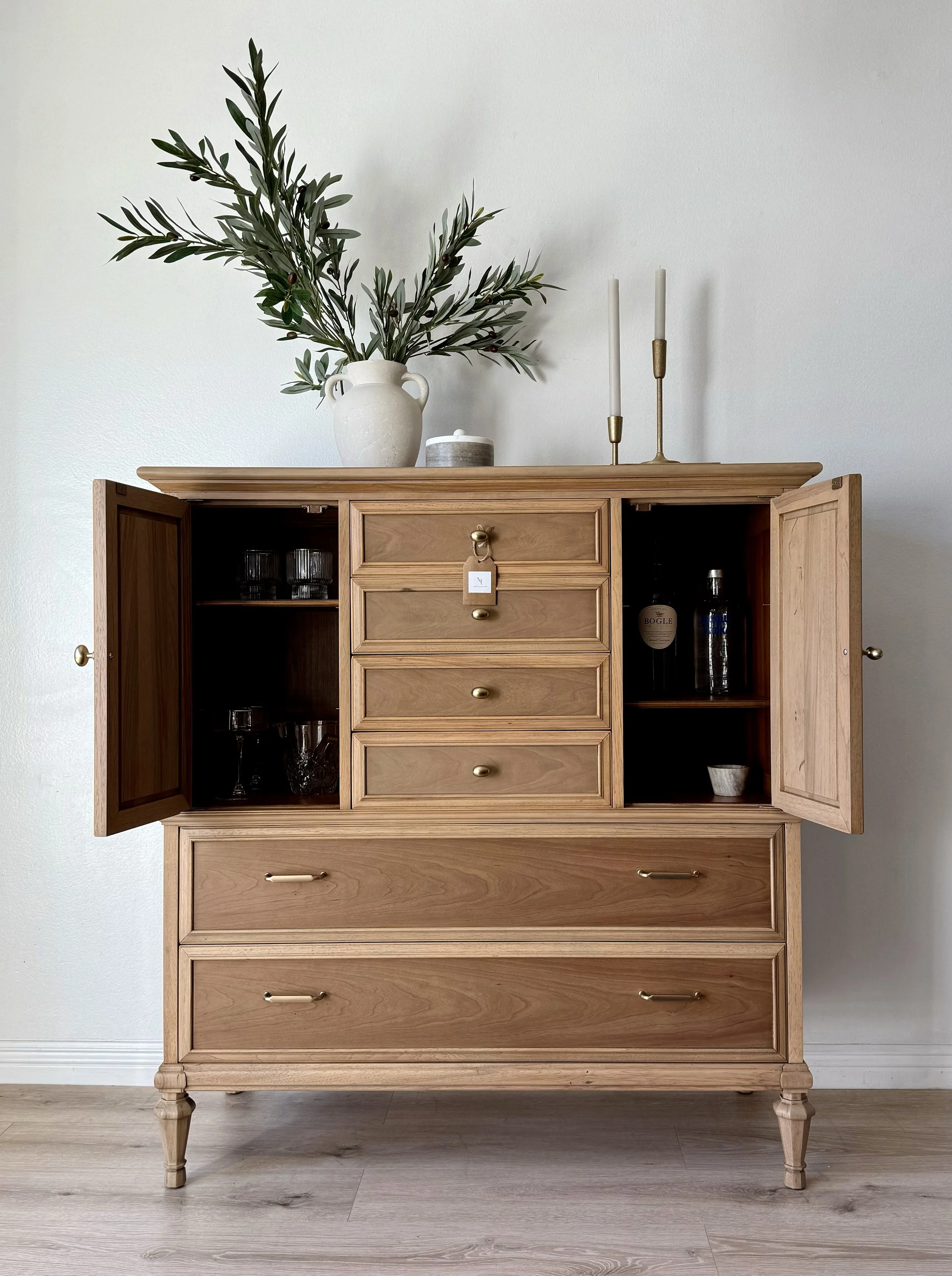 A wooden cabinet with open doors revealing glassware and bottles inside, topped with a large white vase holding green branches, two tall candle holders, and a small container, set against a plain white wall.