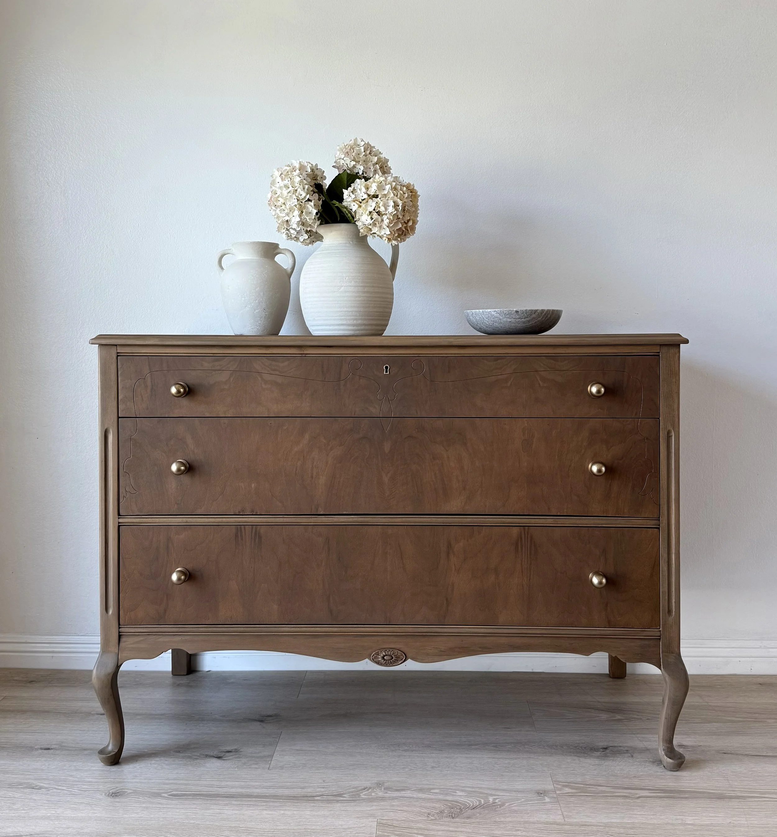 A wooden dresser with three drawers and curved legs, decorated with three vases and a bowl on top, set against a plain white wall.