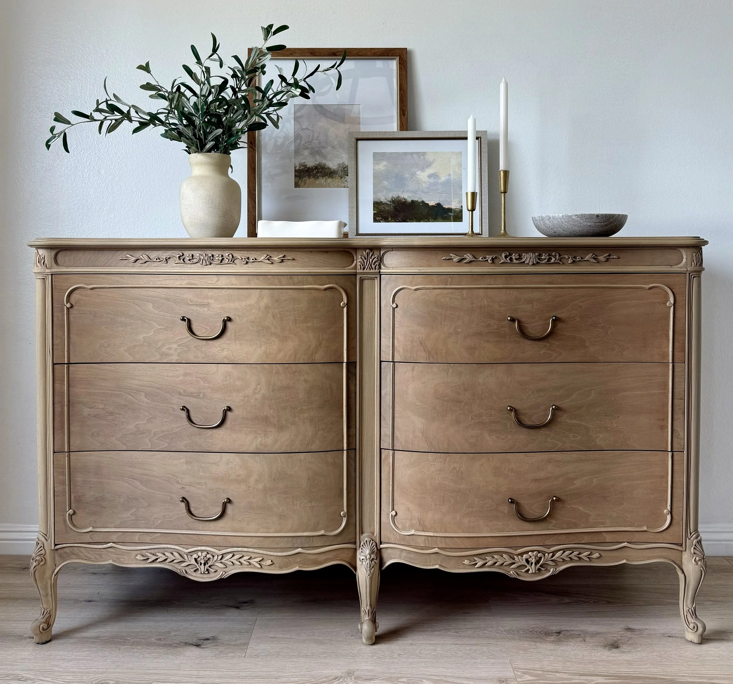 A vintage wooden dresser with ornate carvings, topped with a large vase of greenery, framed artworks, candlesticks, and a bowl, against a plain white wall.