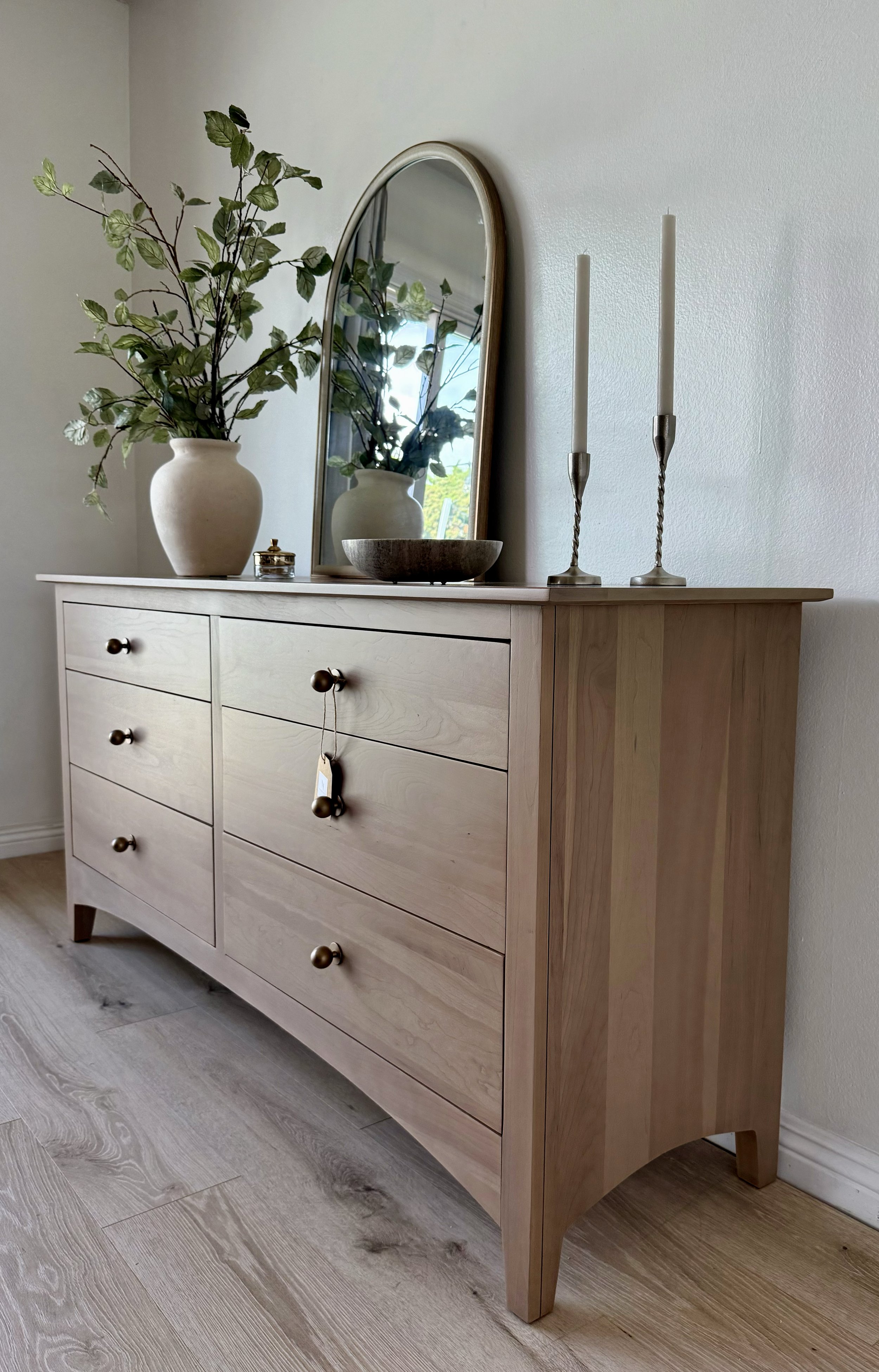 A wood dresser with six drawers, a oval mirror, a vase with green leaves, a bowl, and two tall candleholders with white candles.