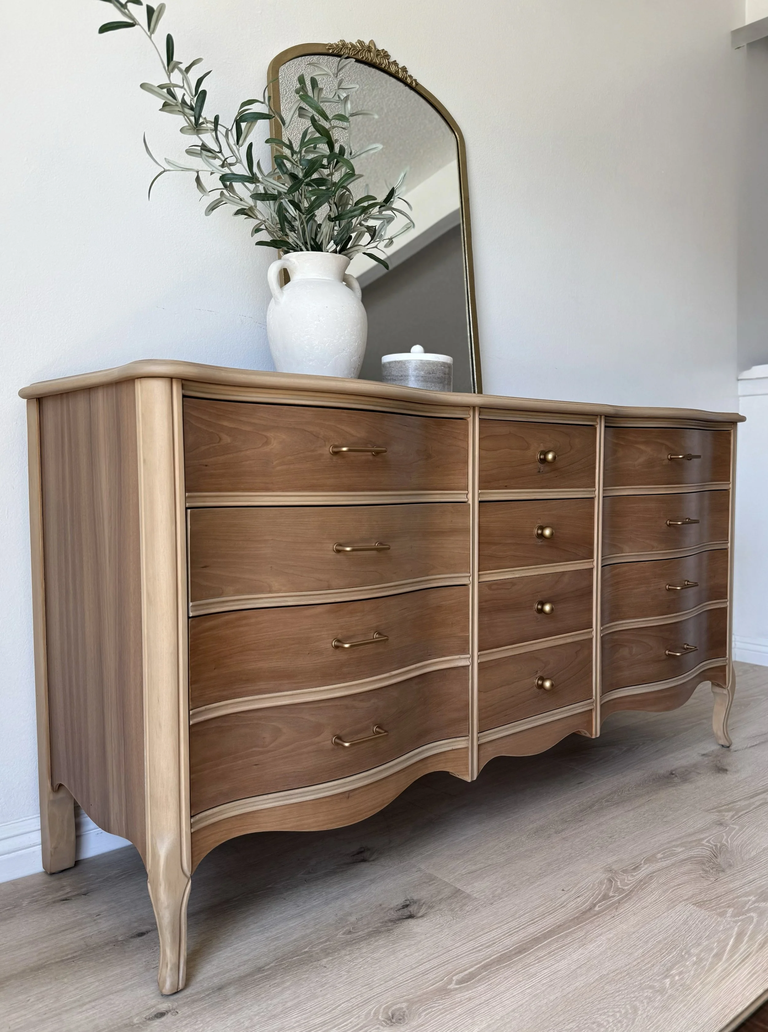 A wooden dresser with brass handles and wavy drawer fronts, topped with a white vase holding green leafy branches, a small round container, and a tall mirror reflecting part of the room.