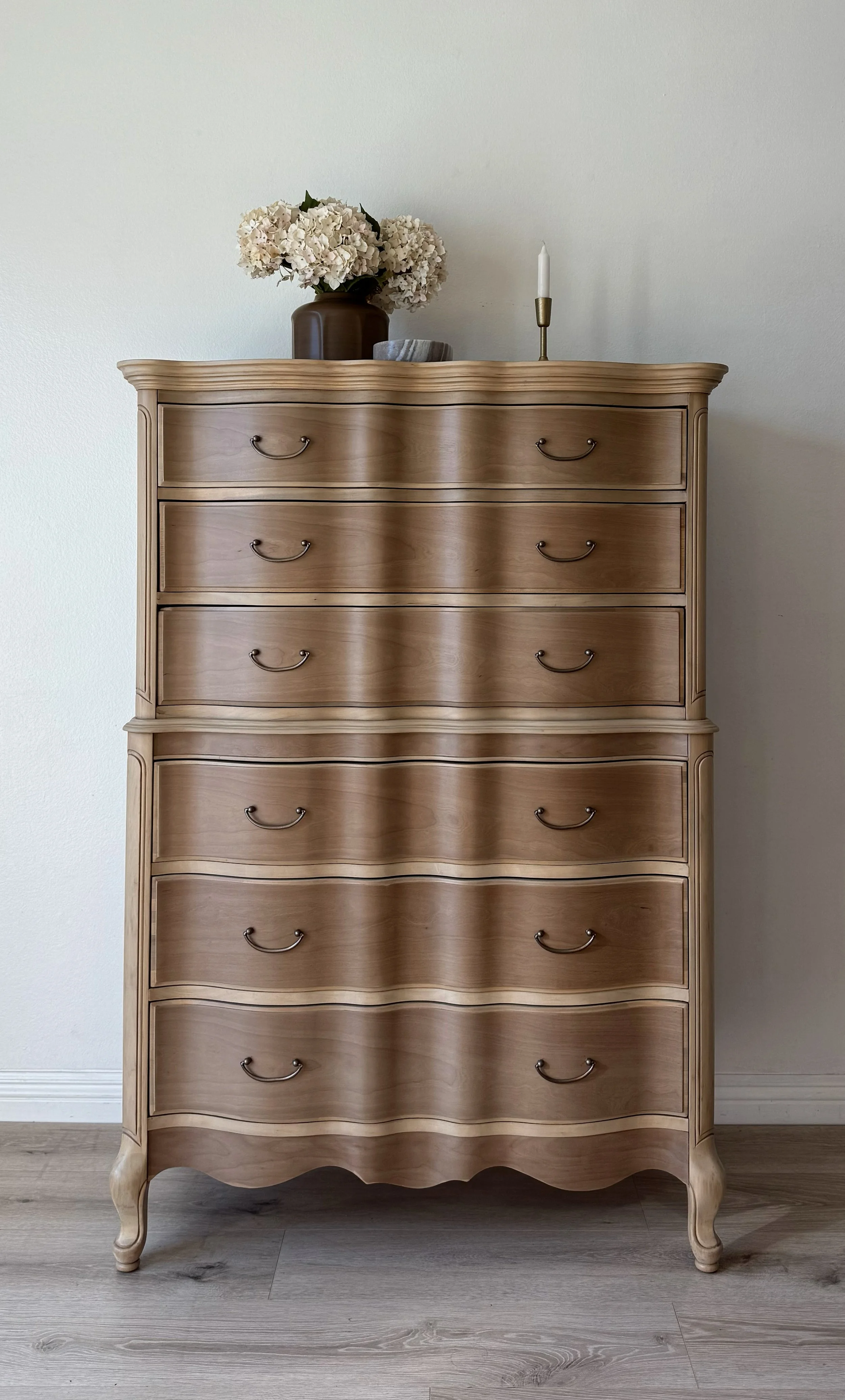 A wooden dresser with five drawers, decorated with a vase of white flowers, a candle, and a decorative bowl on top.