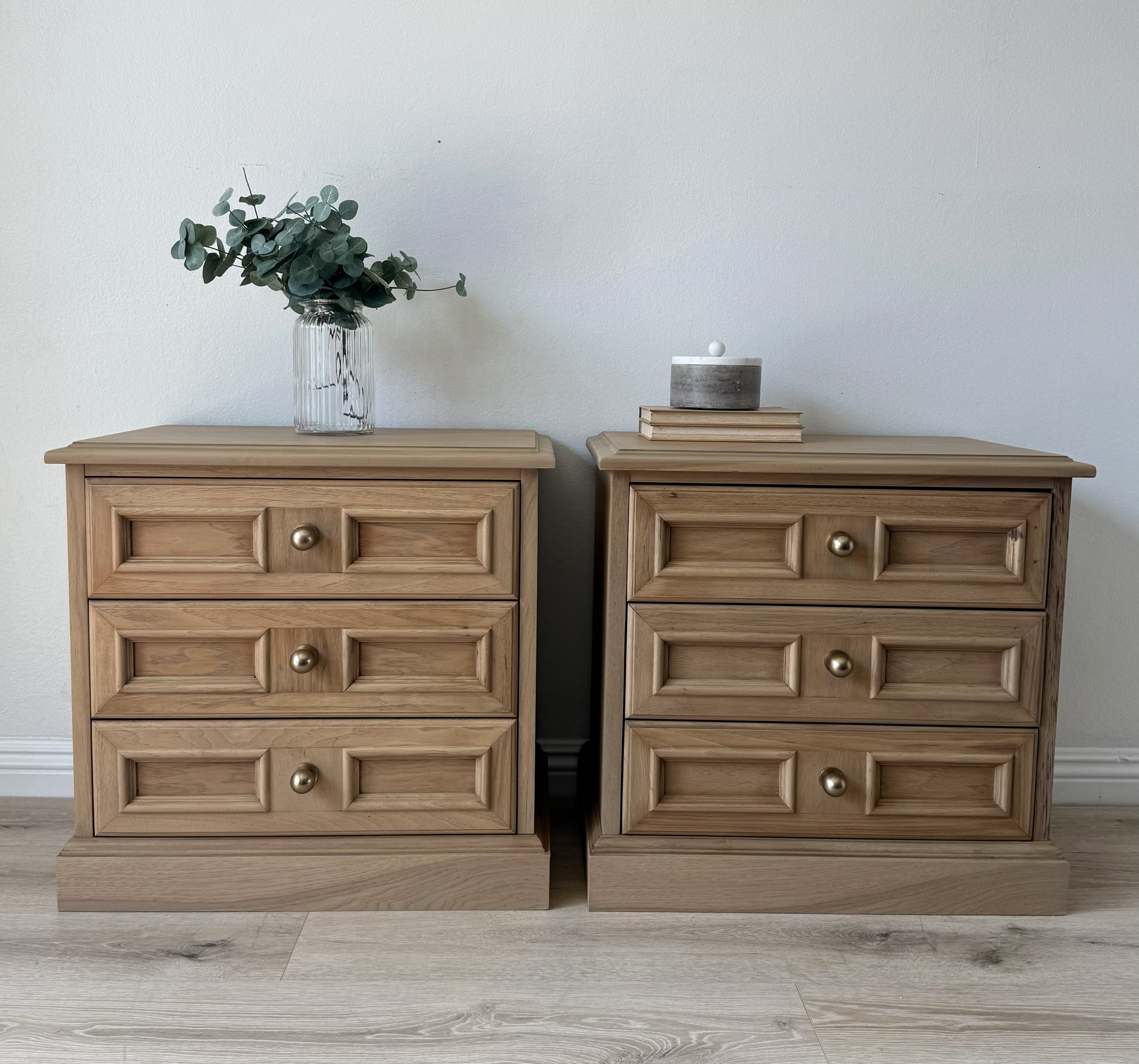 Two wooden nightstands with three drawers each, topped with a vase of greenery on the left and stacked books with decorative object on the right, against a white wall.