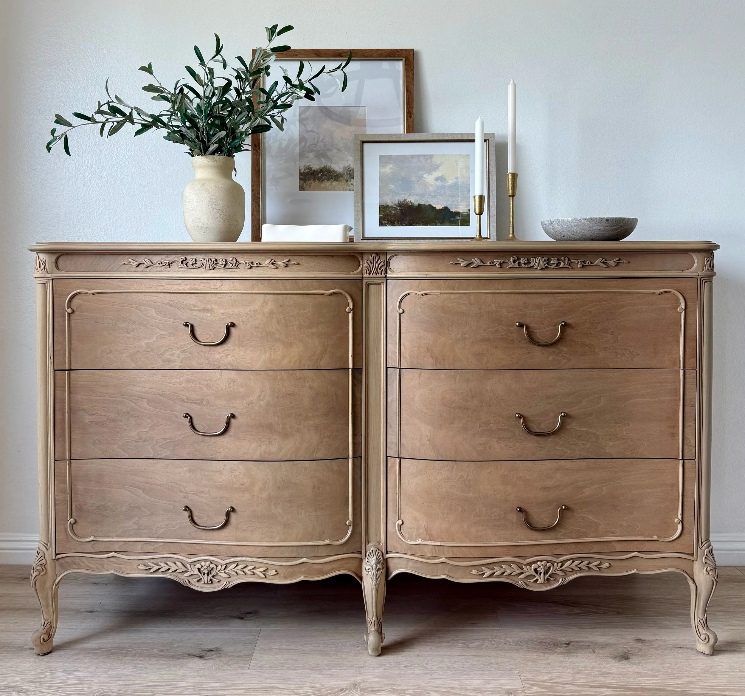 A vintage wooden dresser with intricate carvings, decorated with a vase of greenery, framed artwork, and candlesticks, against a white wall.