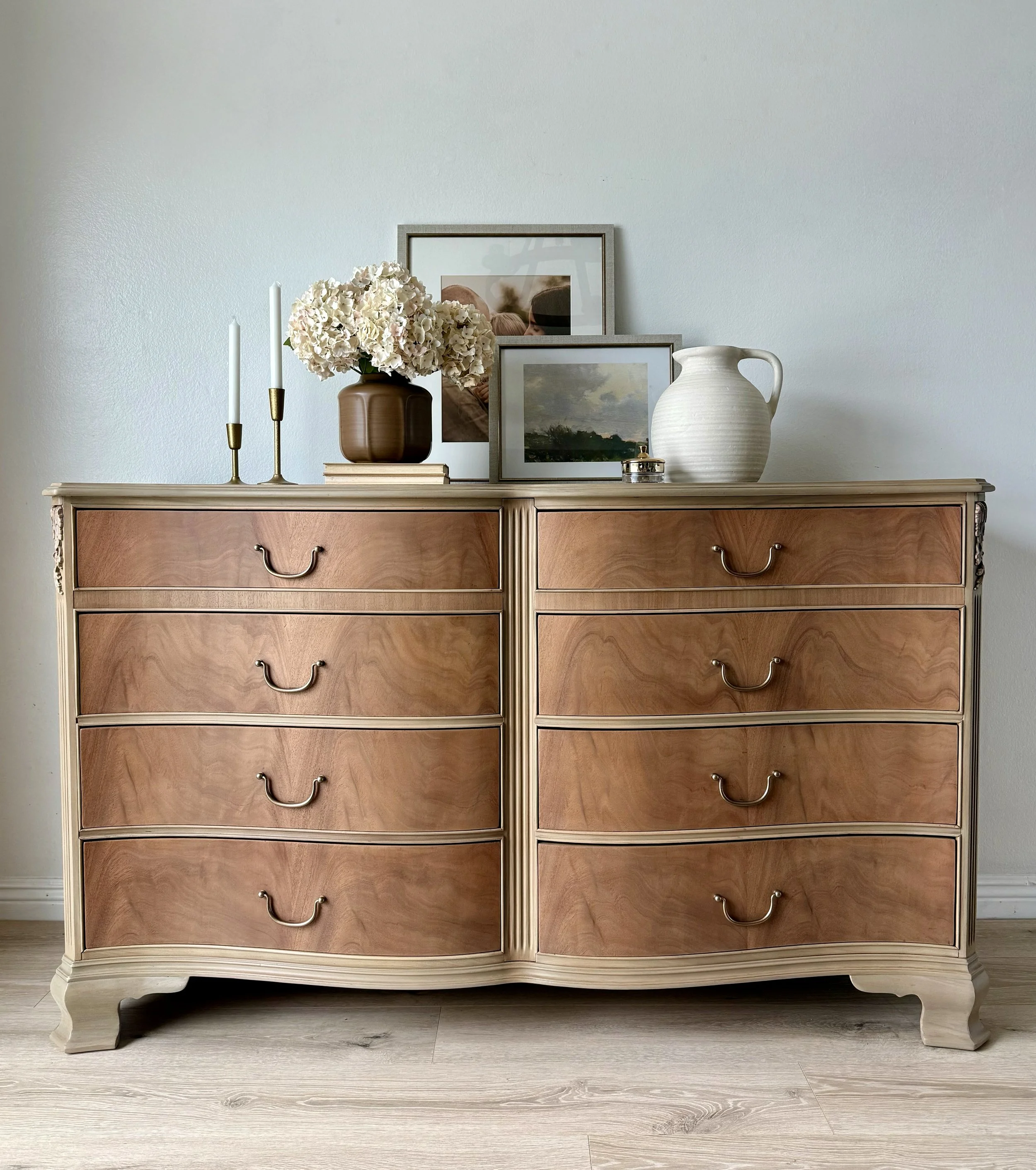 A vintage wooden dresser with six drawers, adorned with a vase of white flowers, framed pictures, a white pitcher, and candlesticks on top.