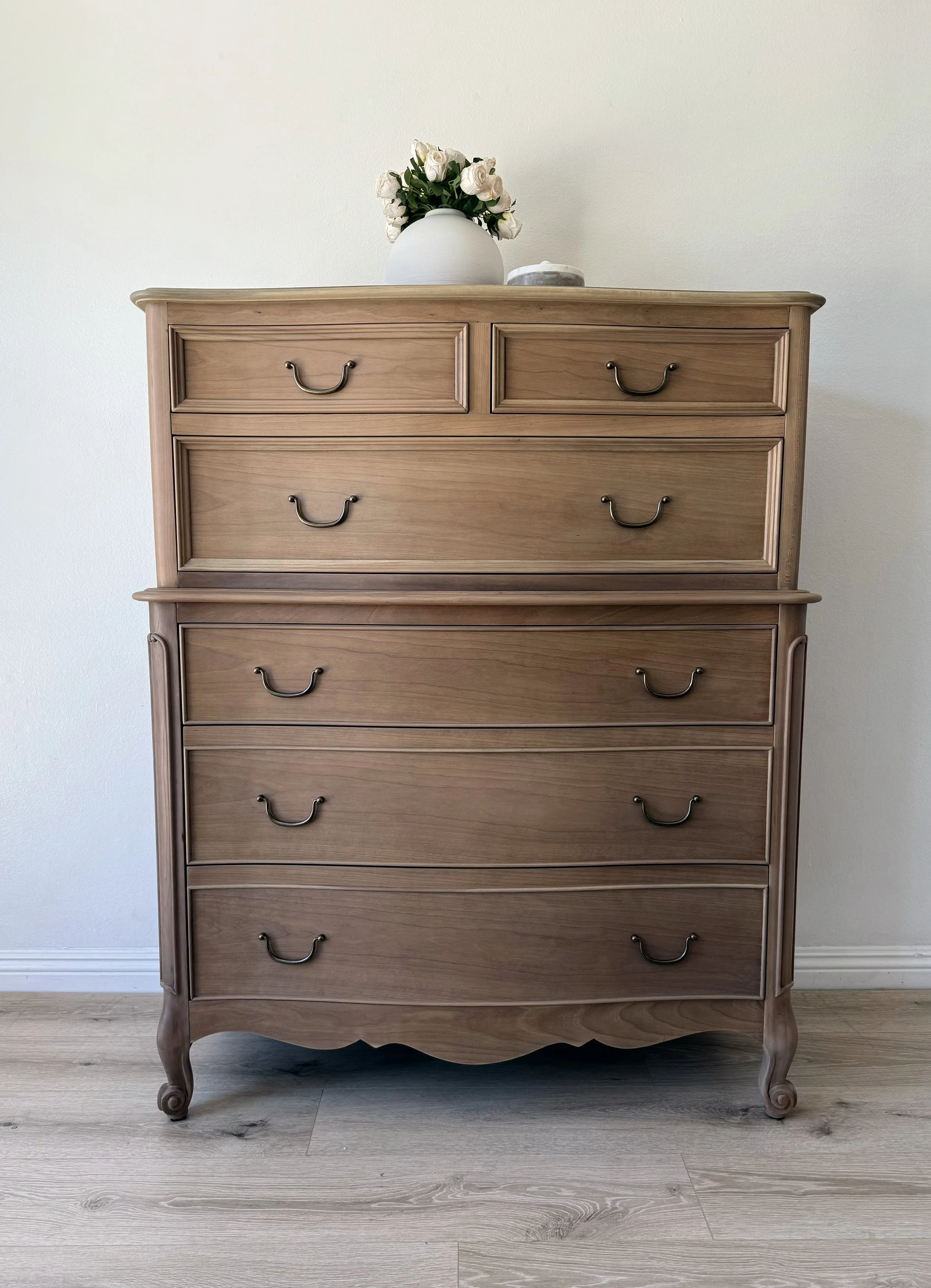 Wooden dresser with five drawers and metallic handles, topped with a white vase of white roses and a small container, set against a plain white wall.