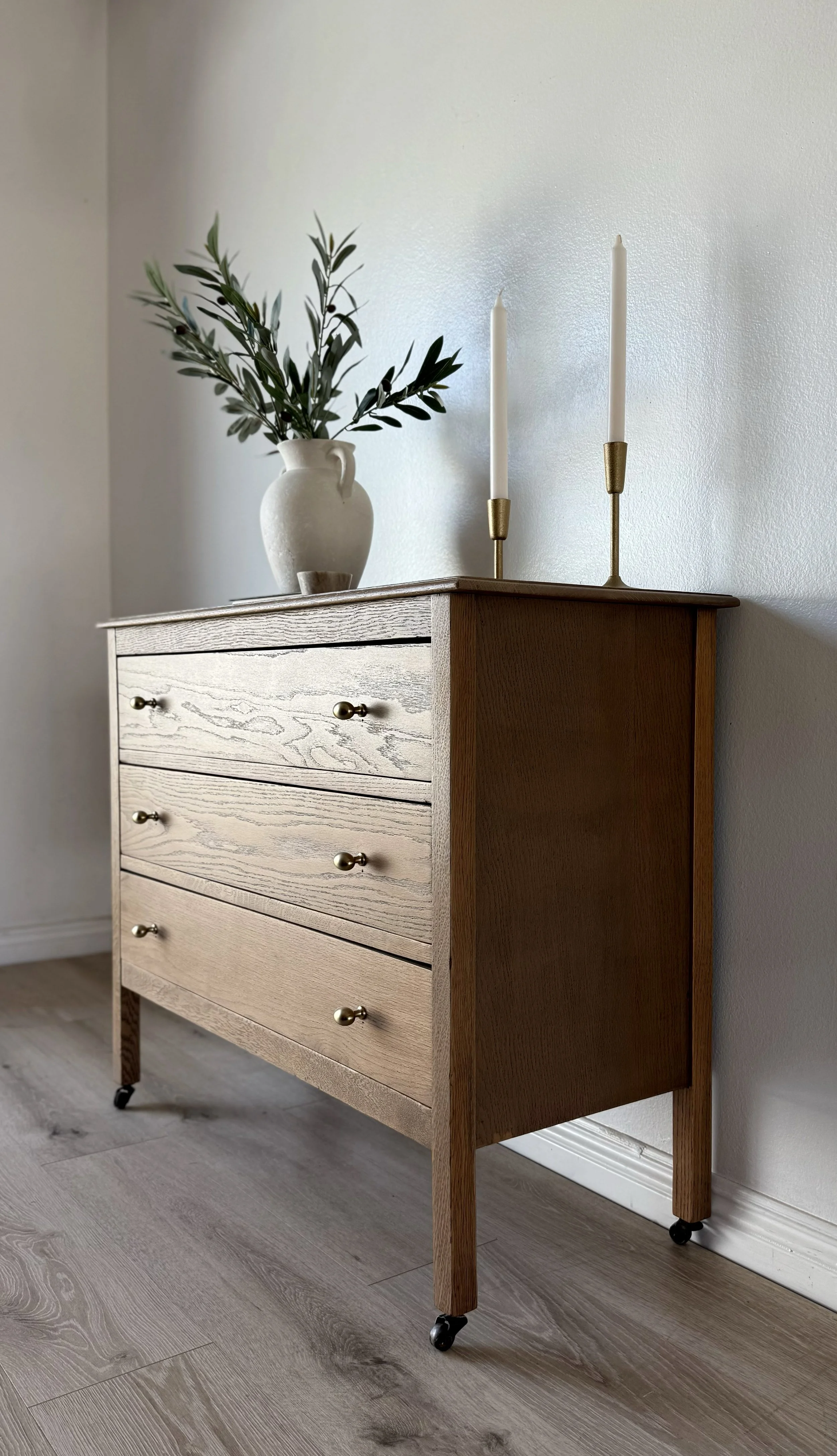 Wooden dresser with four drawers, brass knobs, on wheels, topped with a white vase with greenery and two white candles on gold candlesticks.