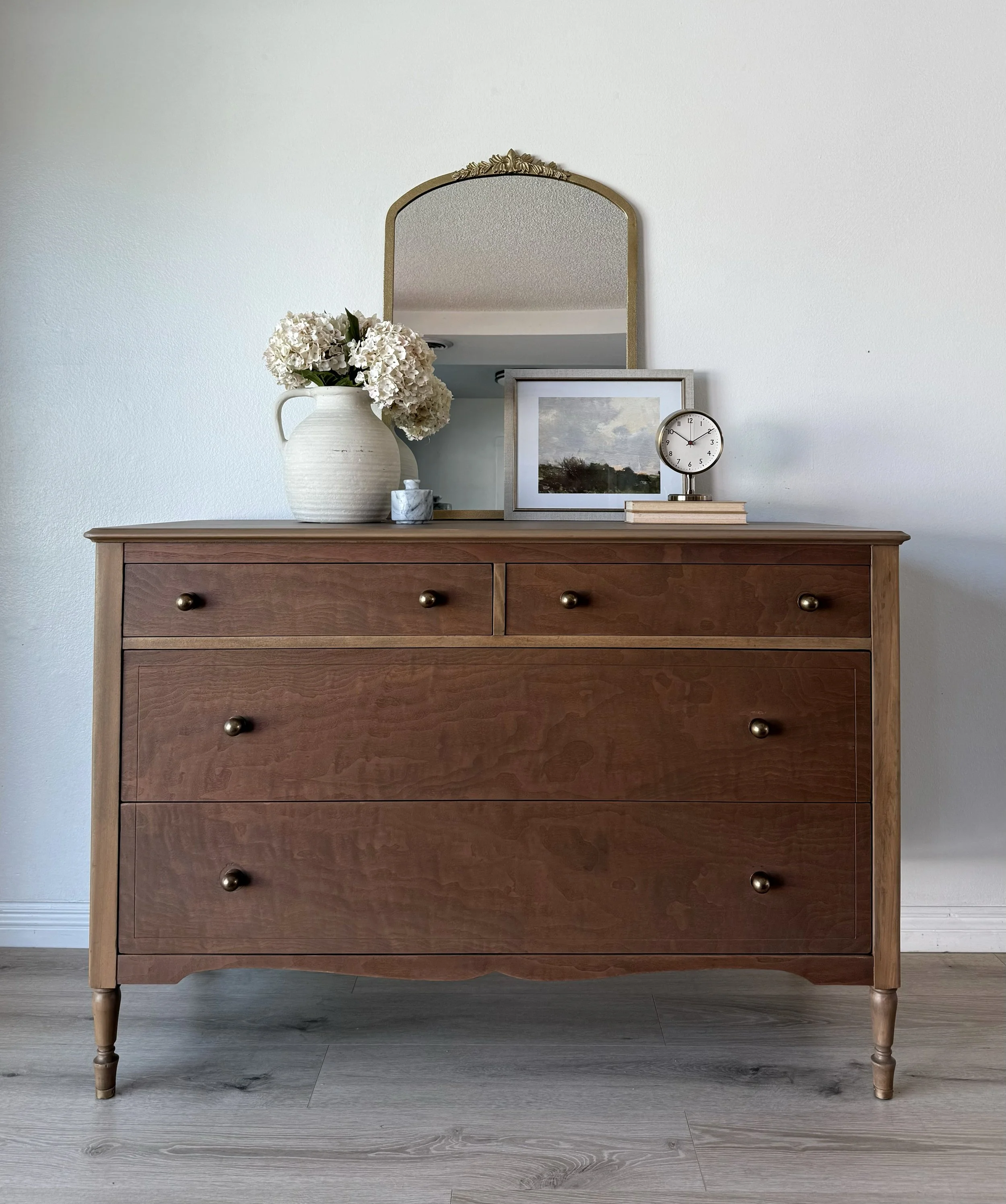 Wooden dresser with a mirror, floral vase, framed photo, clock, and stacked books on top.