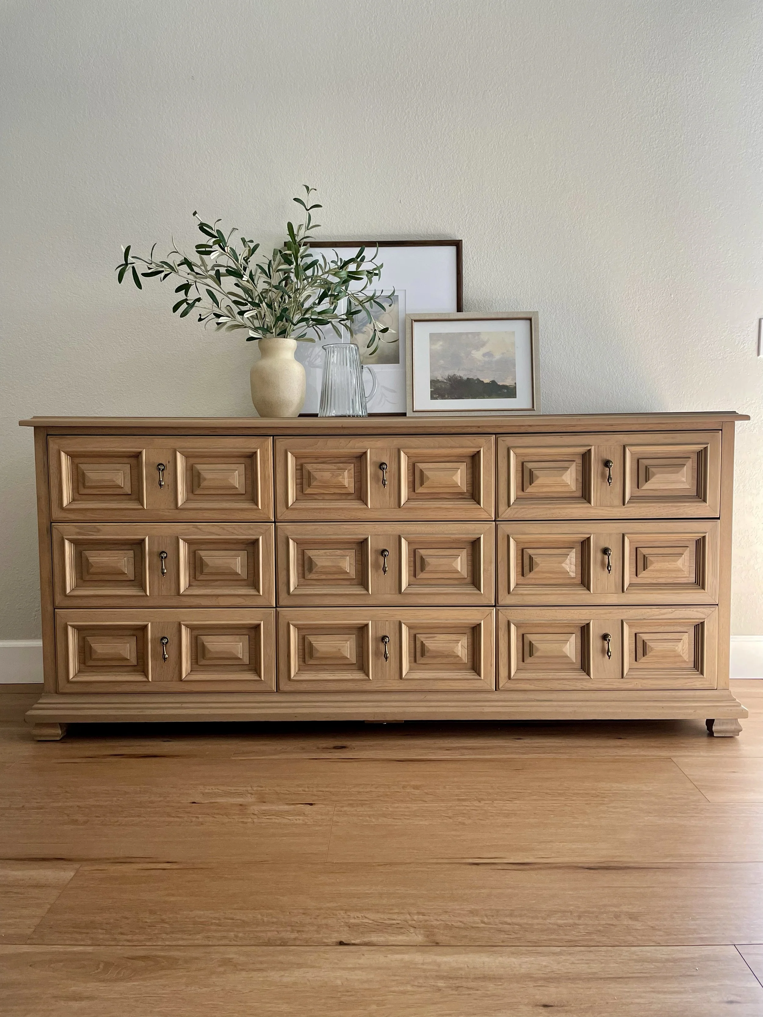 Wooden dresser with geometric carved patterns on drawer fronts, topped with a vase of olive branches, a glass pitcher, and framed artwork, against a plain wall.