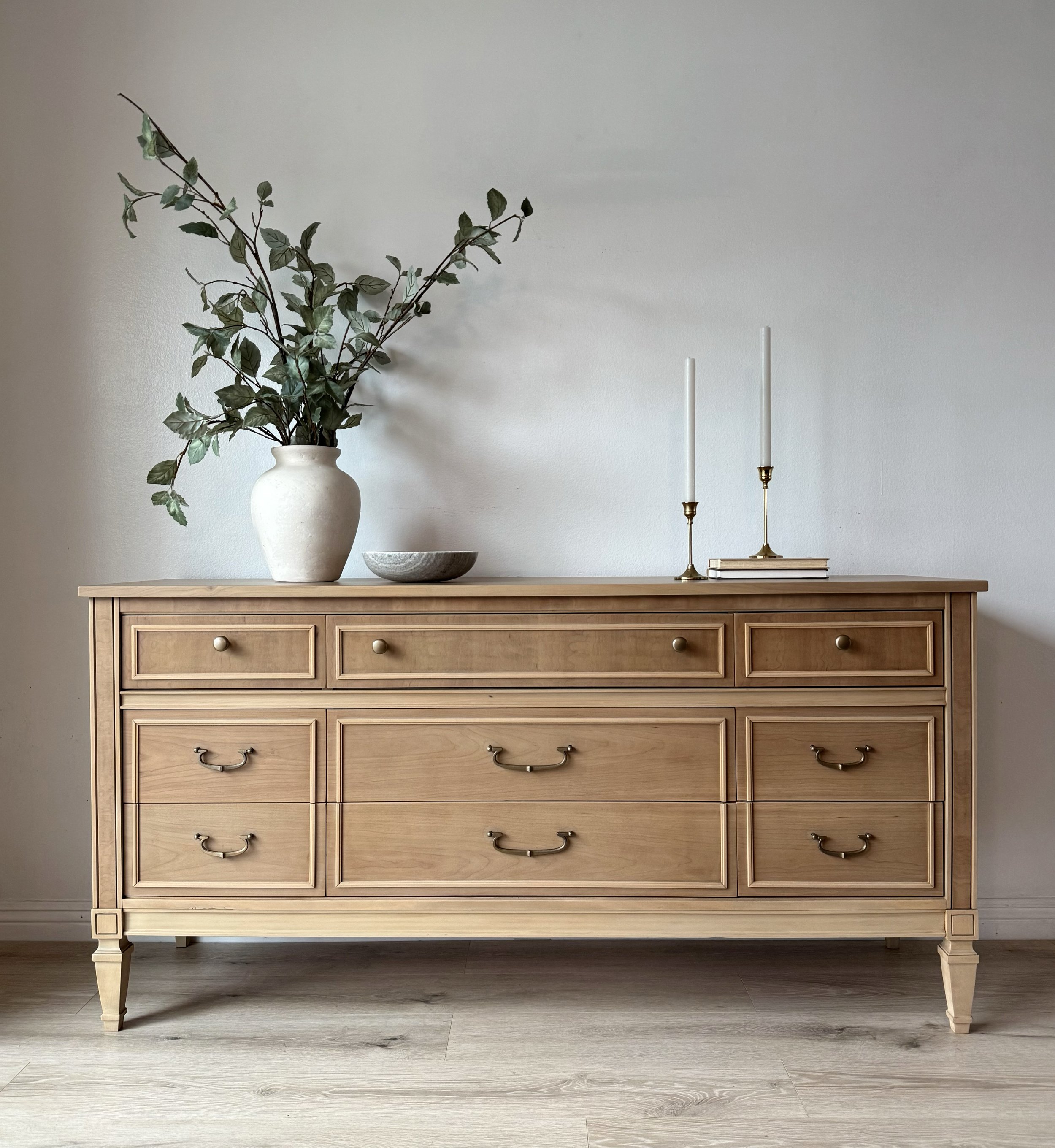A wooden dresser with a large white vase containing green leafy branches, a small ceramic bowl, two candlesticks with unlit candles, and a stack of books on top, against a plain wall.