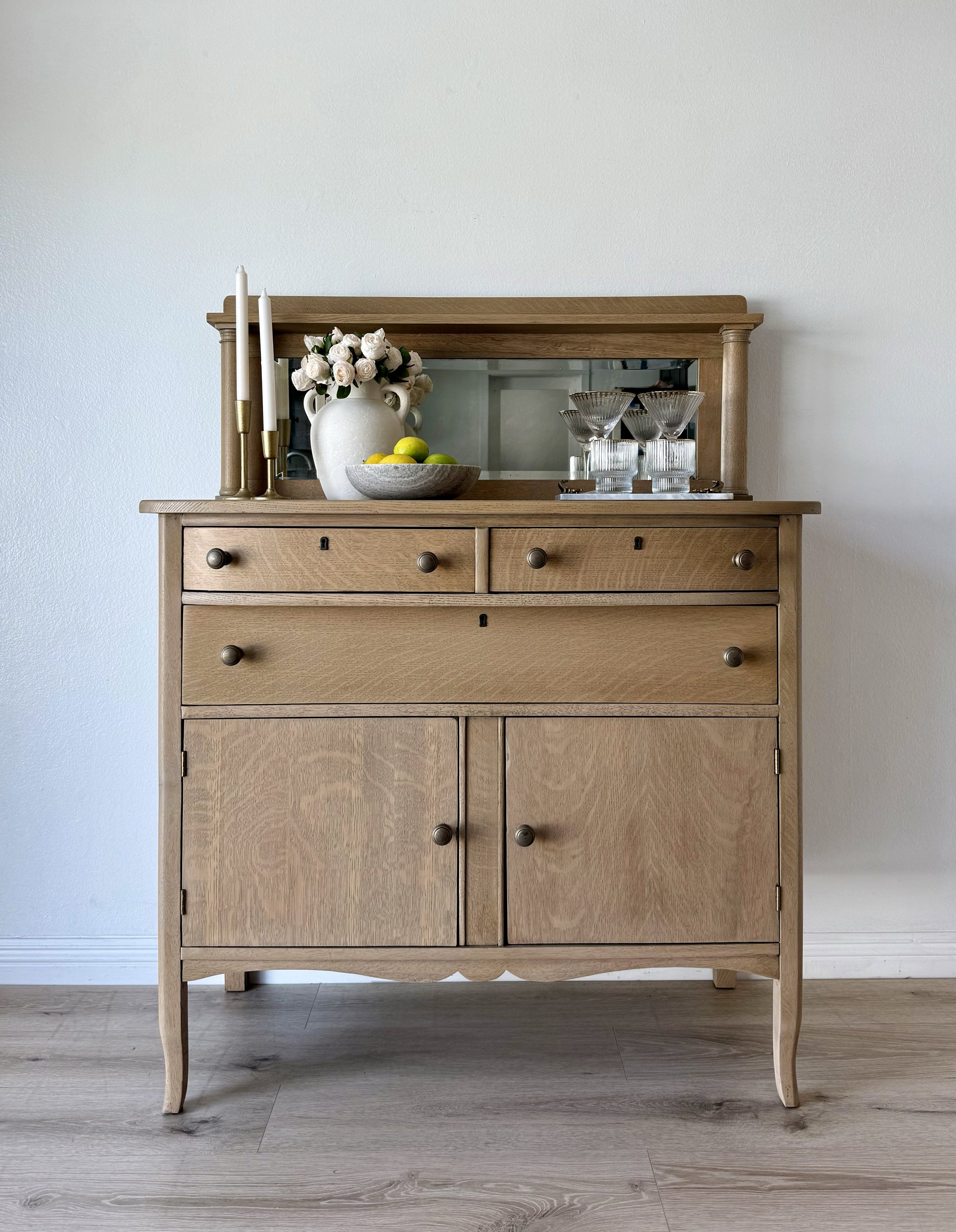 A wooden sideboard with drawers and cabinets, decorated with a white vase of white roses, gold candlesticks with white candles, a bowl of lemons and green apples, and glassware, with a mirror behind it against a plain white wall.