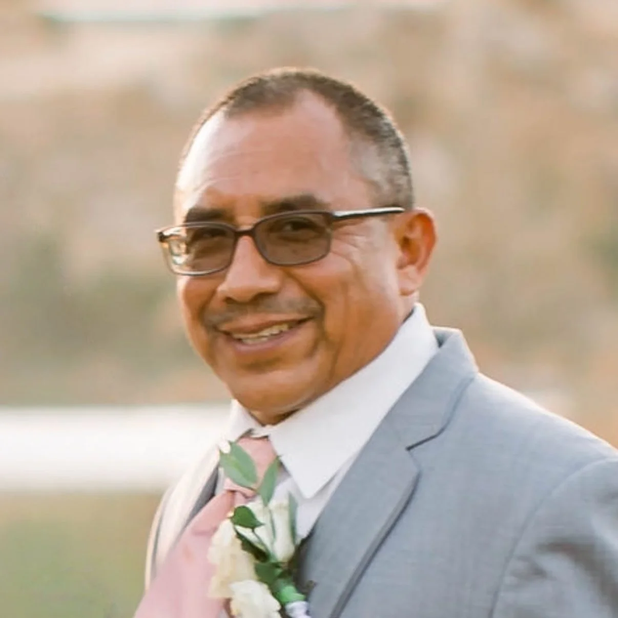 A man smiling outdoors, wearing glasses, a light gray suit, white shirt, and a pink tie with a boutonnière.