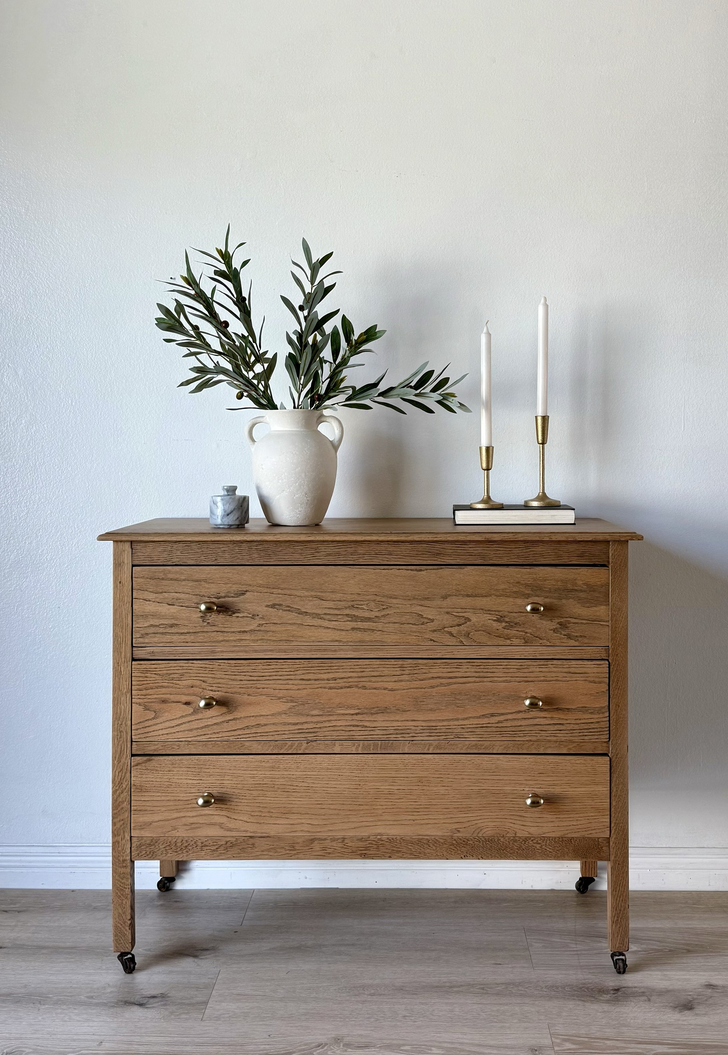 A wooden dresser with three drawers, brass knobs, and small caster wheels, set against a white wall with light wood flooring. Decor items on top include a white vase with green foliage, a small gray marble container, and a stack of books with two whi
