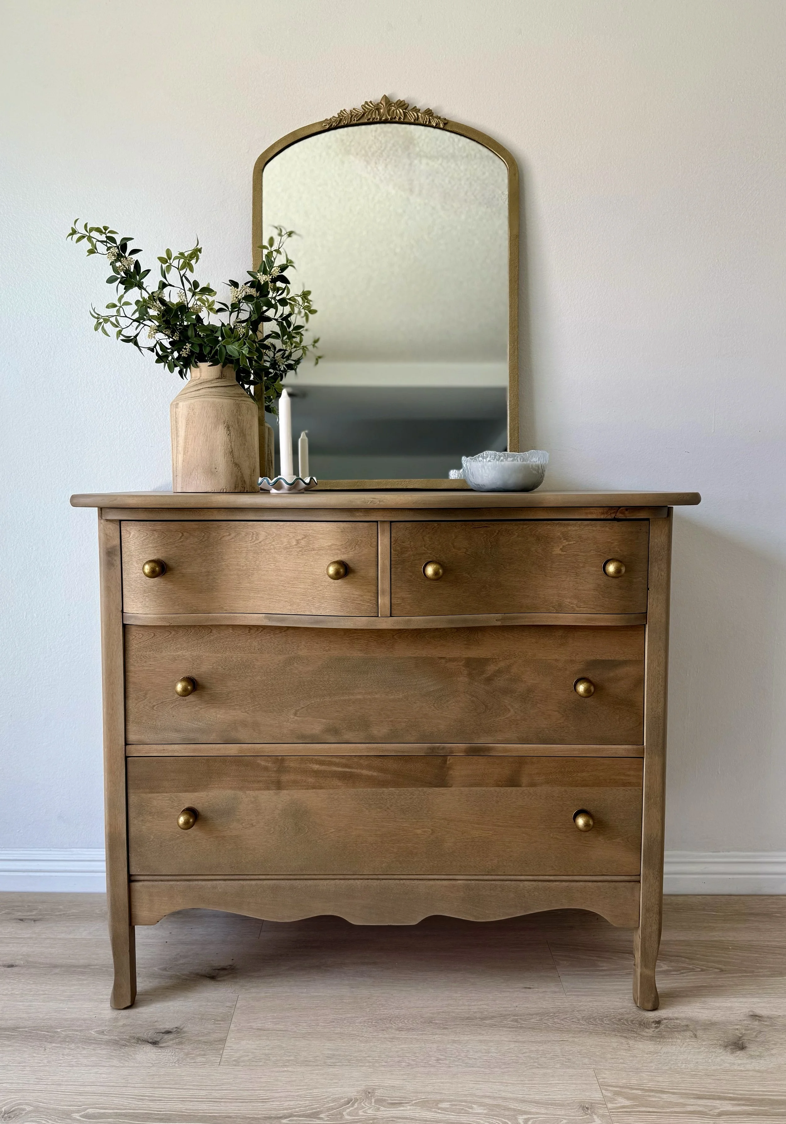 Wooden dresser with gold knobs, topped with a large vase of green foliage, white candles, and a ceramic bowl, reflected in a tall mirror with gold trim, set against a plain white wall.