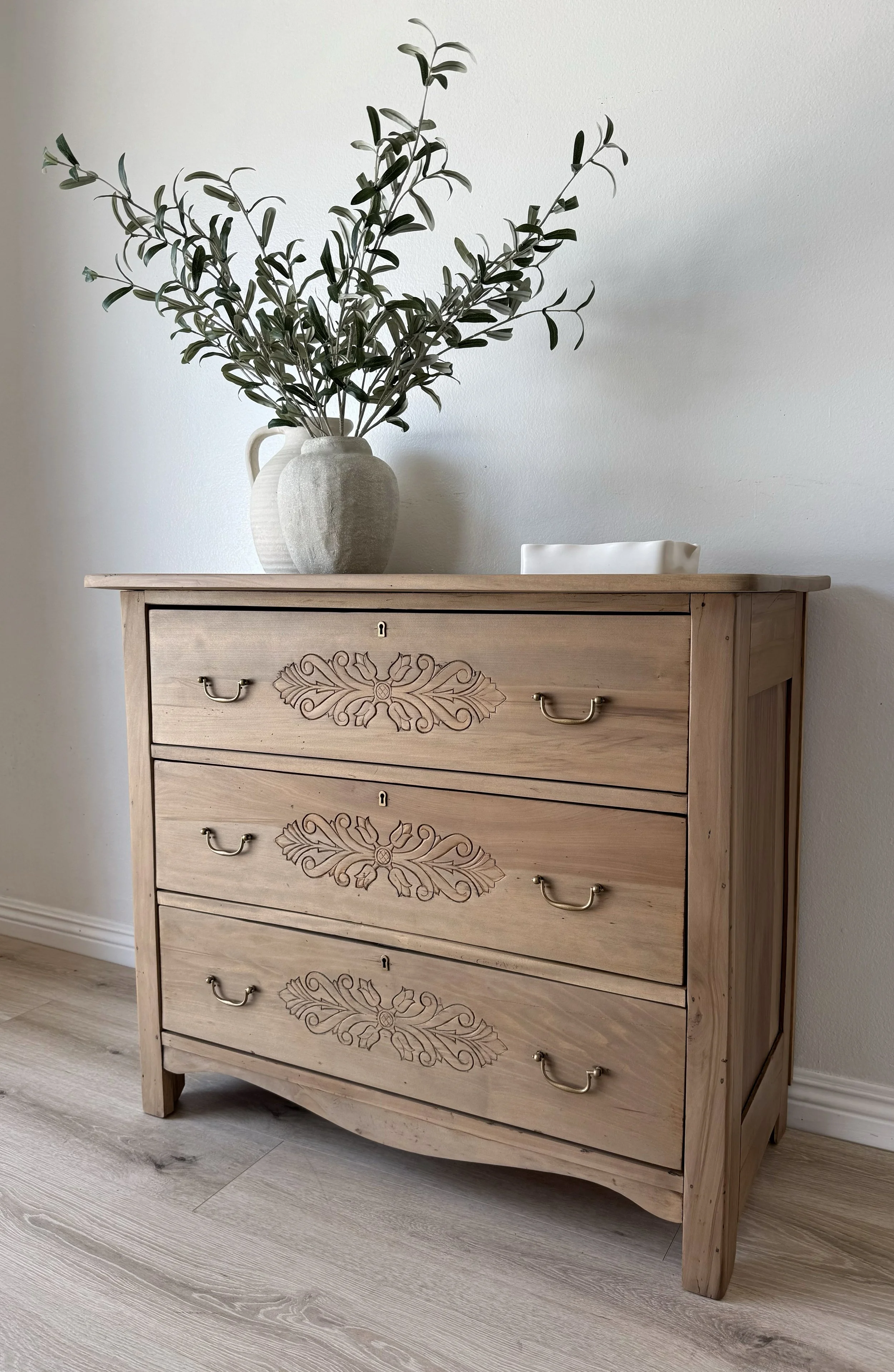 A wooden dresser with three drawers, each adorned with decorative floral carvings and brass handles, placed in front of a white wall. On top of the dresser, there are two ceramic vases, one tall and the other shorter, with greenery in the taller vase