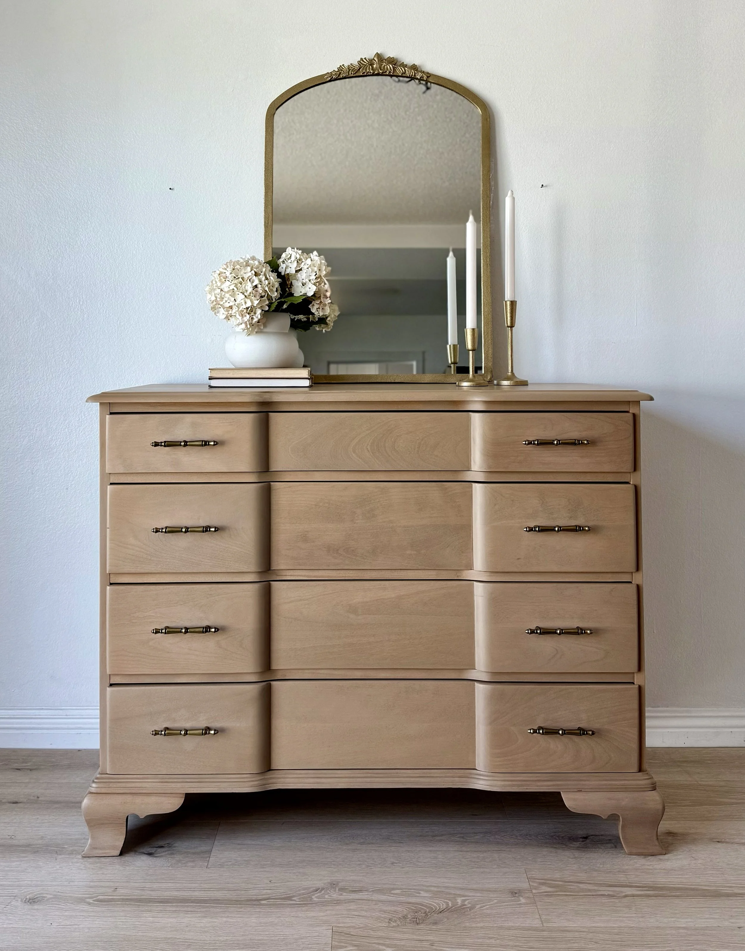 A wooden dresser with six drawers, topped with a mirror, a white vase with white flowers, some books, and three white candles in gold candle holders.