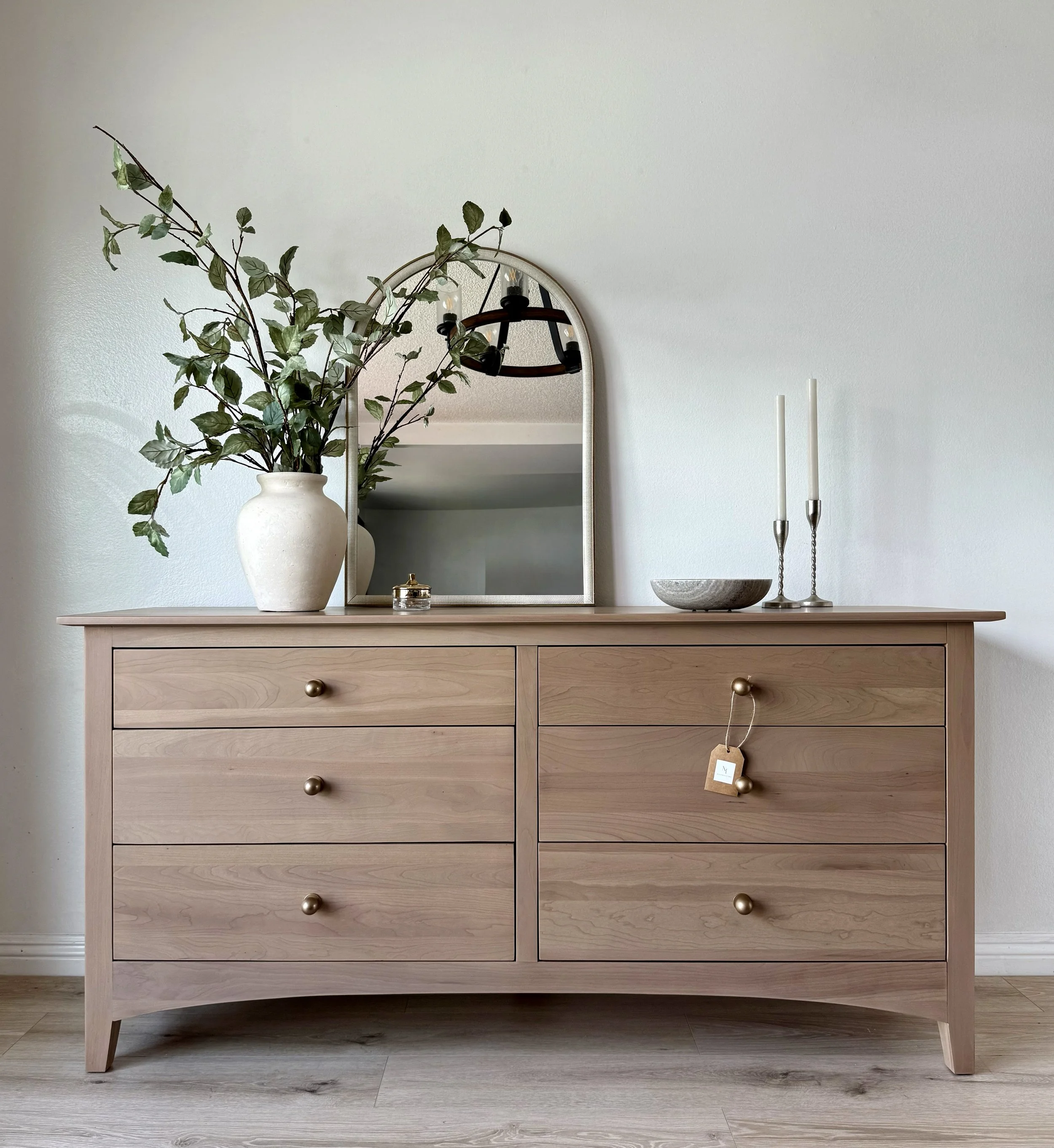 Wooden dresser with brass knobs, decorated with a white vase holding green branches, a mirror, two white candles in candlesticks, a bowl, and a small jewelry box.