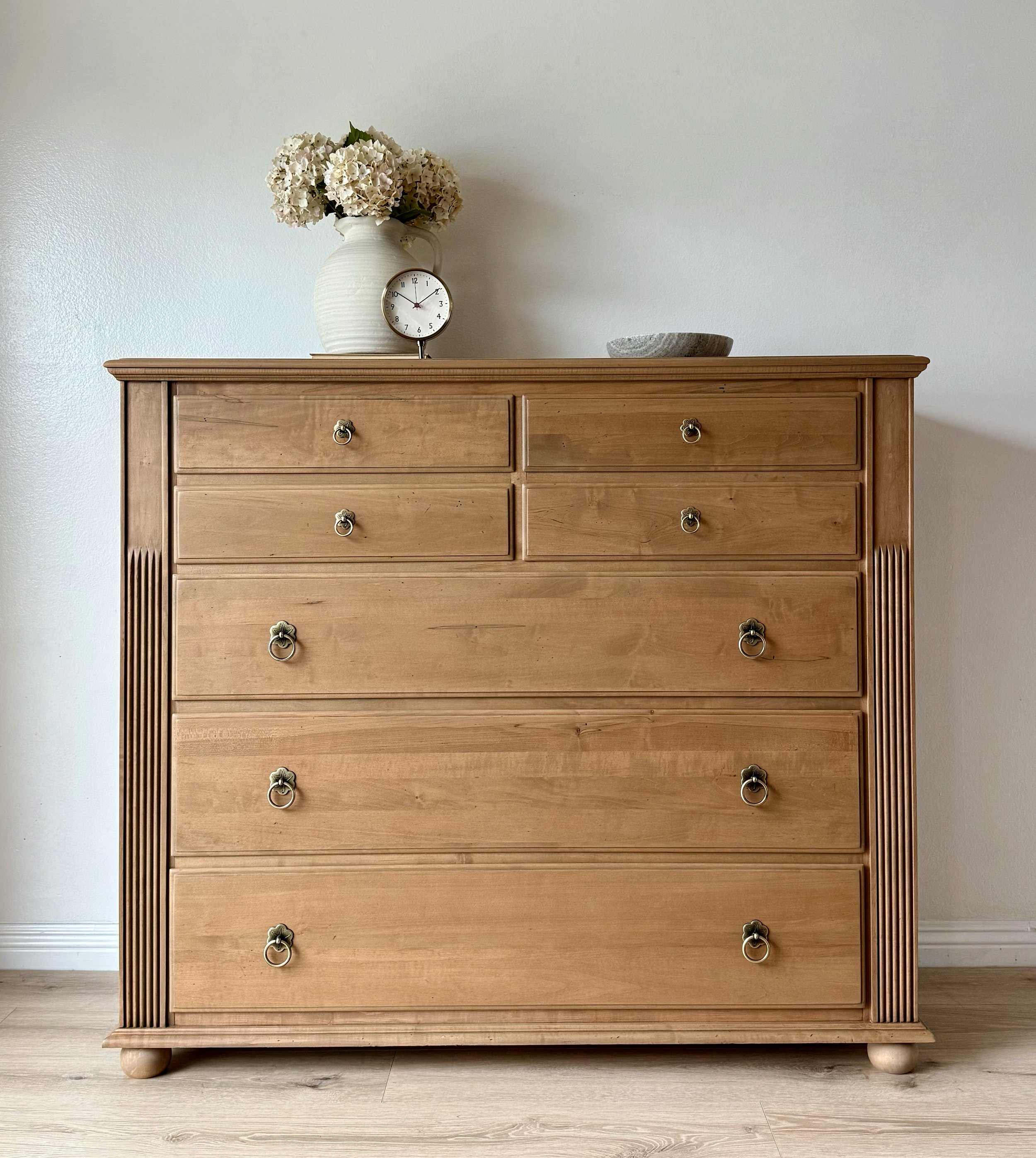 Wooden dresser with six drawers, silver ring pulls, fluted corners, positioned against a plain white wall. Top decor includes a white vase with hydrangeas, a small clock, and a gray tray.