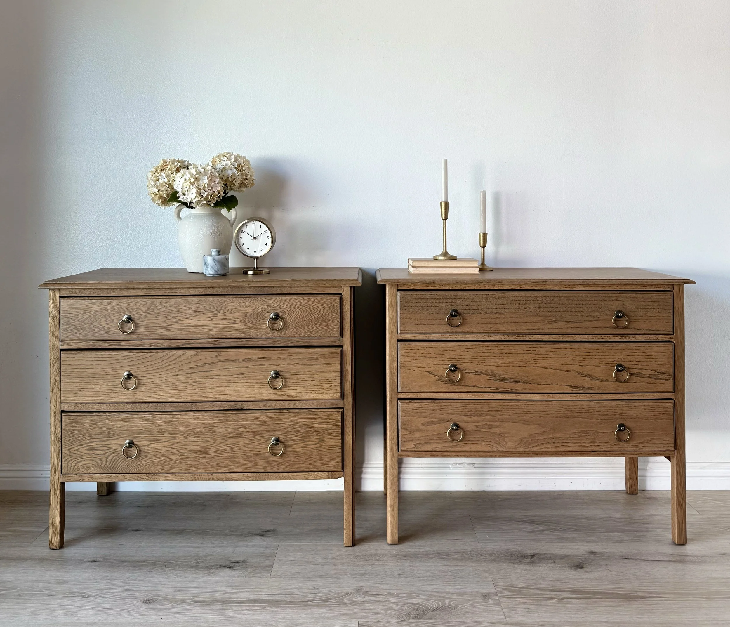 Two wooden dressers placed side by side against a white wall. The left dresser has a vase of white flowers, a clock, and a small decorative item on top. The right dresser has two candlesticks with white candles and a small stack of books.