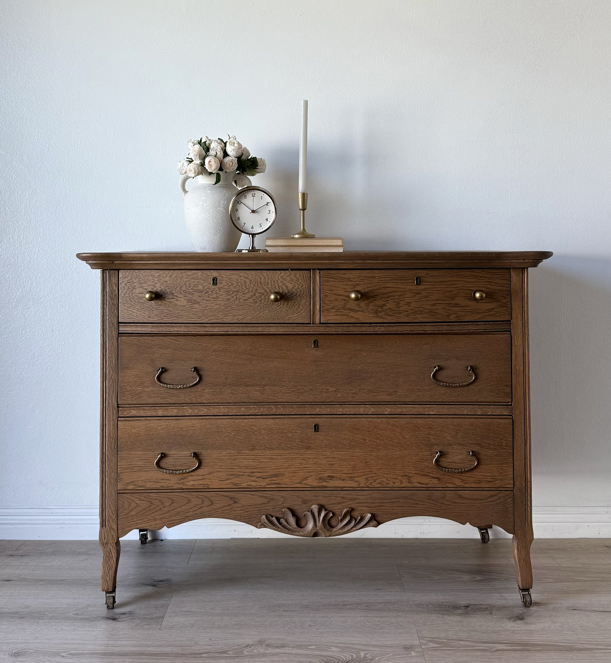 A wooden dresser with a vase of white roses, a clock, a candlestick, and two books on top, against a plain white wall.