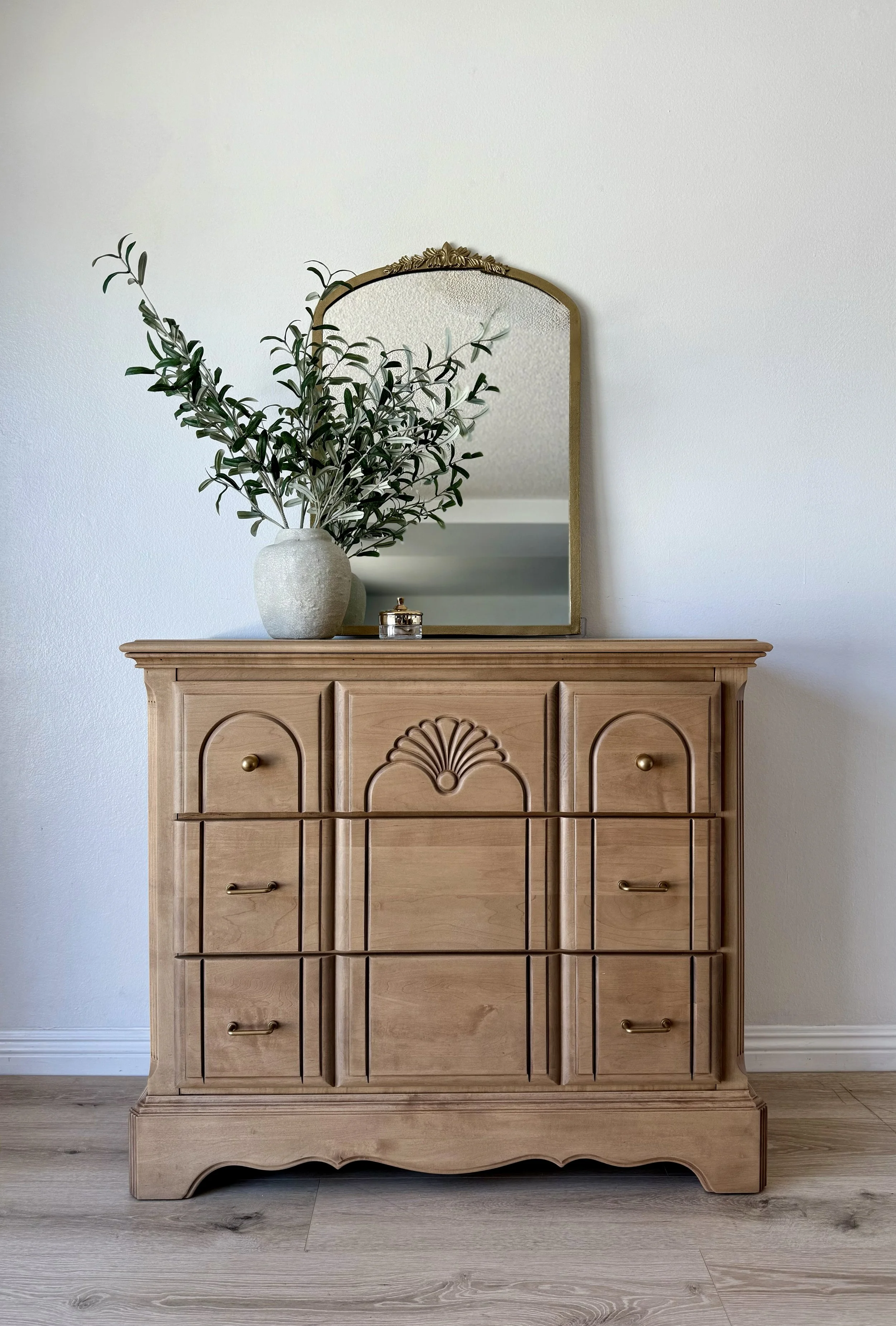 A wooden dresser with decorative carved accents and brass handles, displaying a large vase with green foliage, a small jewelry box, and a mirror mounted on the wall behind it.