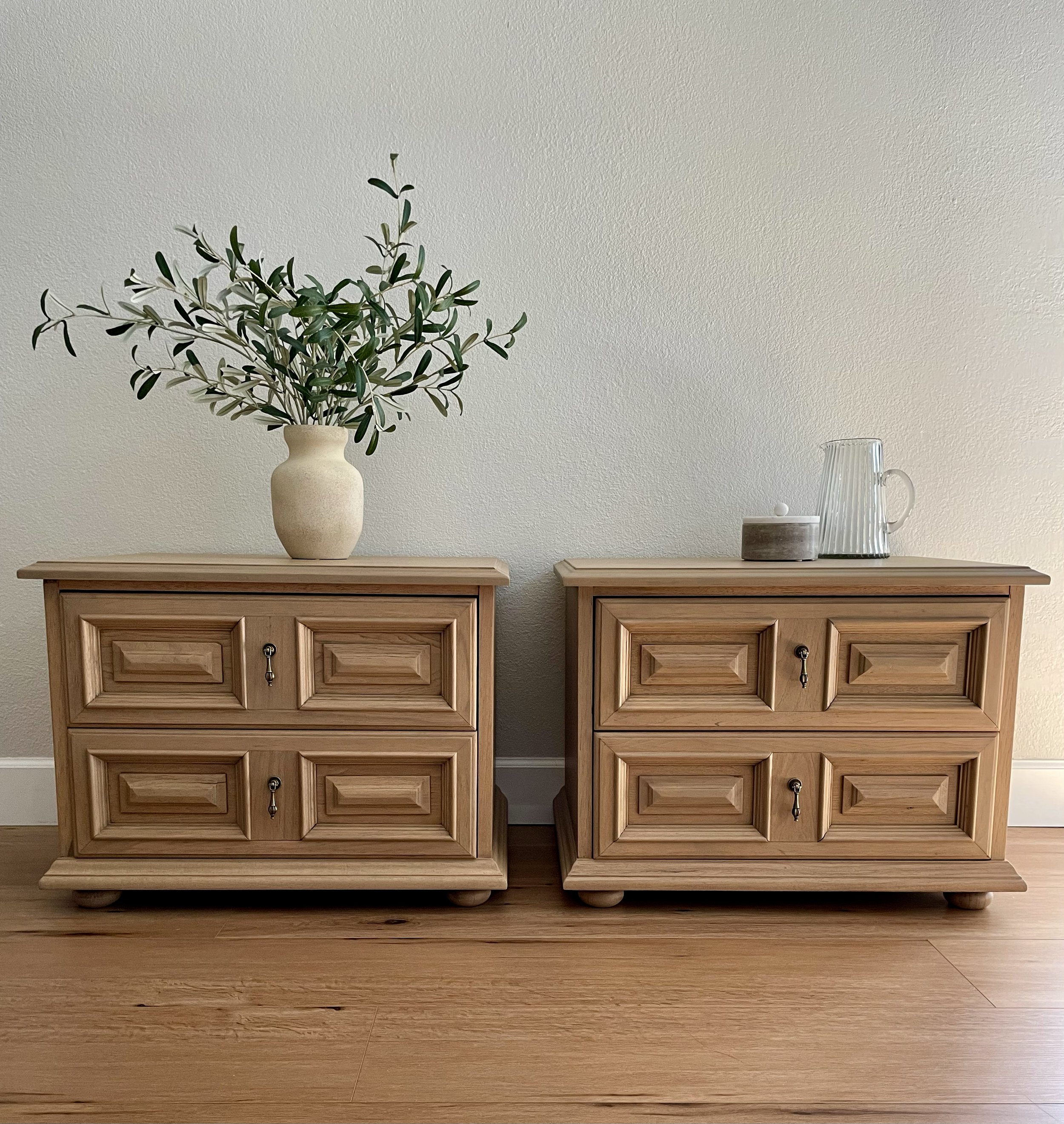 Two wooden nightstands with carved drawer fronts and round feet, each topped with decorative items, against a plain wall and wood floor.